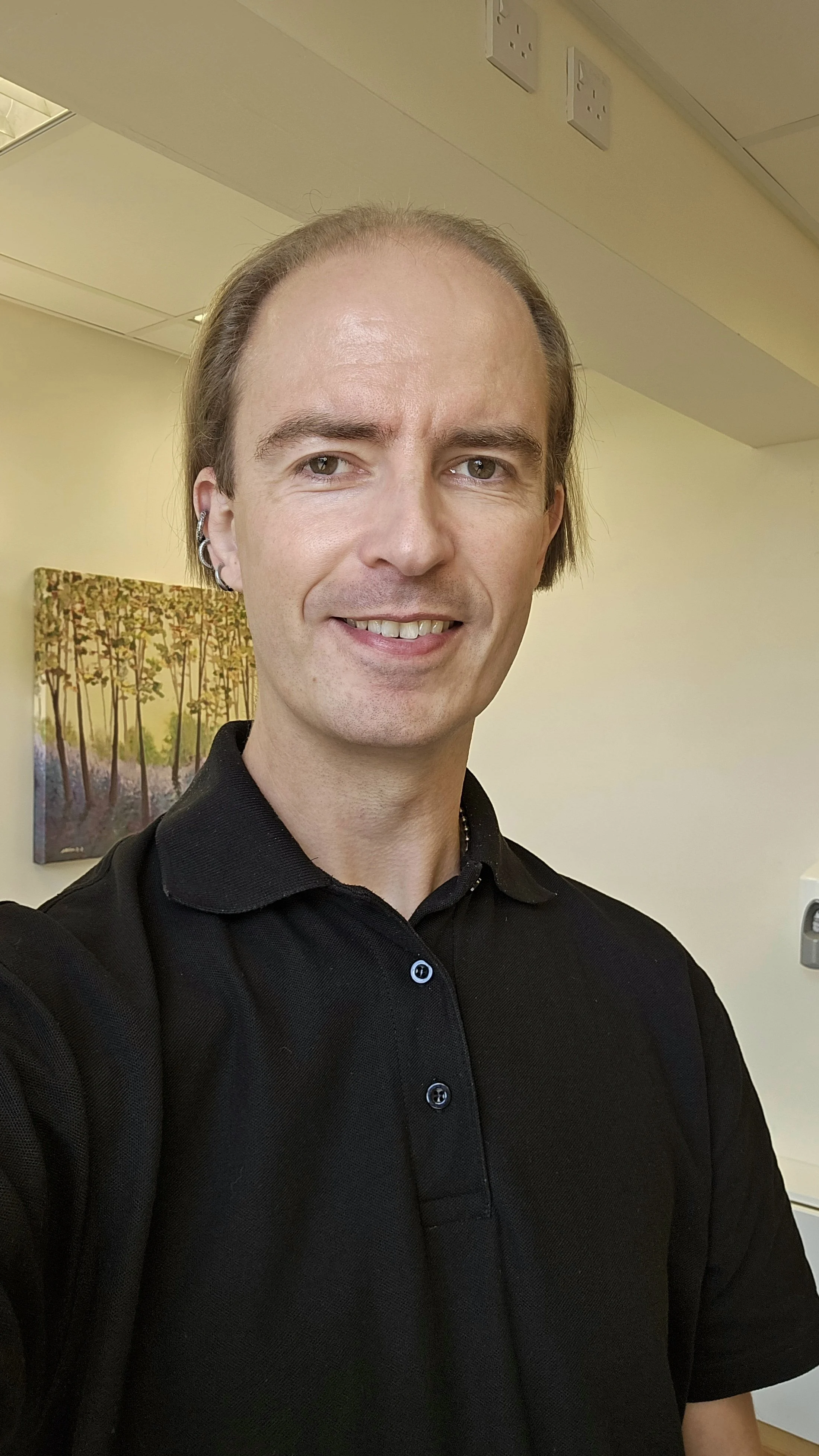 A man with light skin, long brown hair, and multiple earrings takes a selfie indoors, smiling, with a painting of trees on the wall behind him.