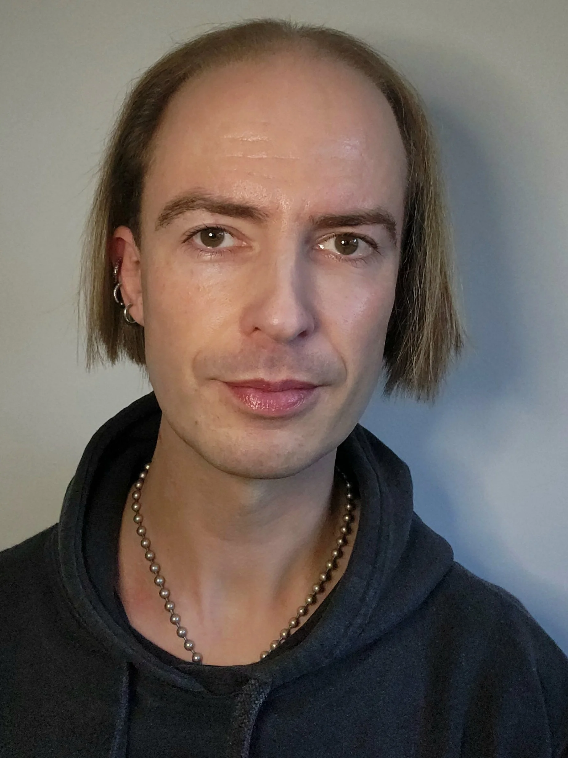 A person with short light brown hair, multiple earrings, wearing a black hoodie and a pearl necklace, standing against a plain background.