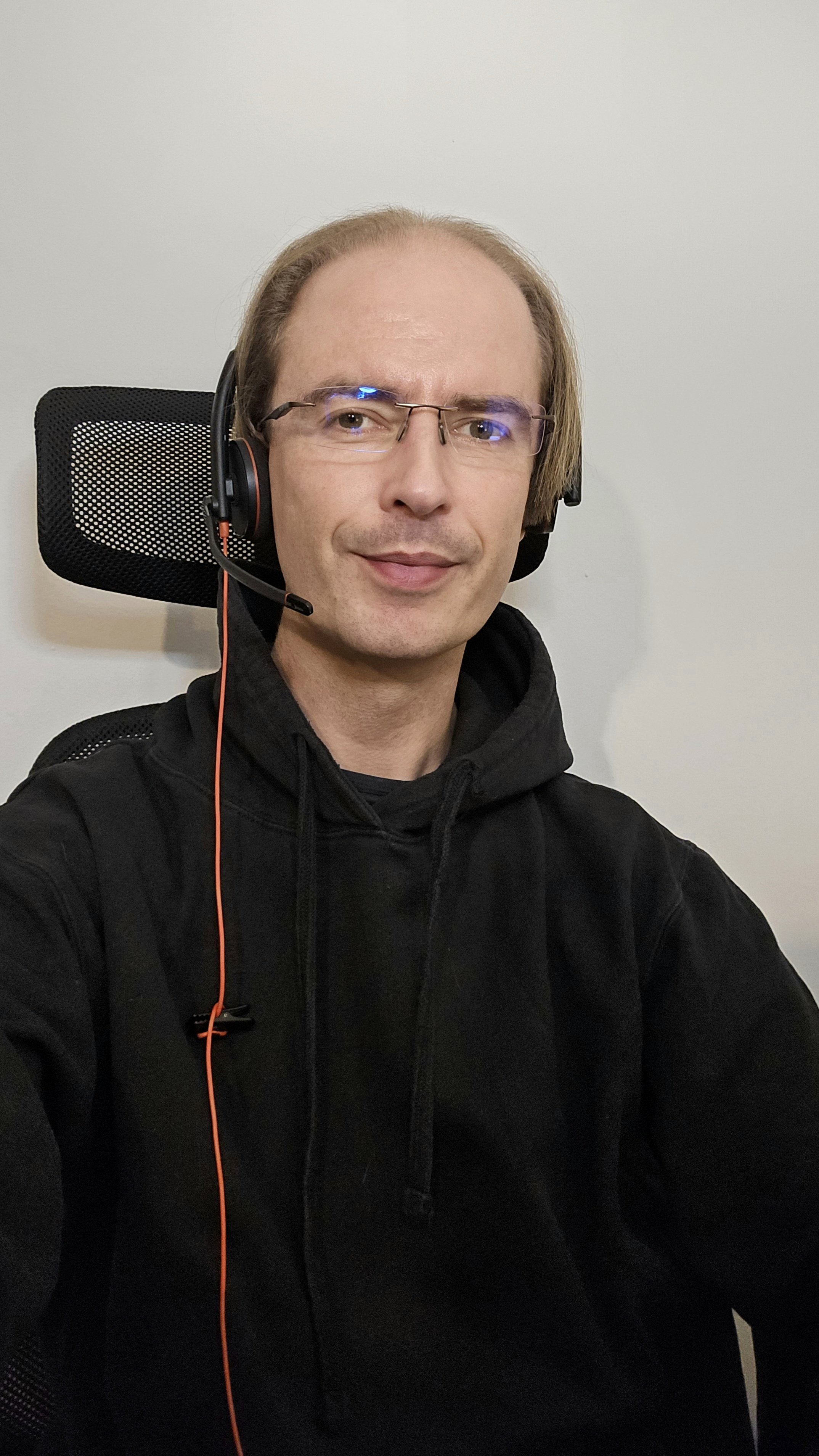 A man with glasses and short, light brown hair wearing a black hoodie and gaming headset, sitting in a black office chair against a plain white wall.