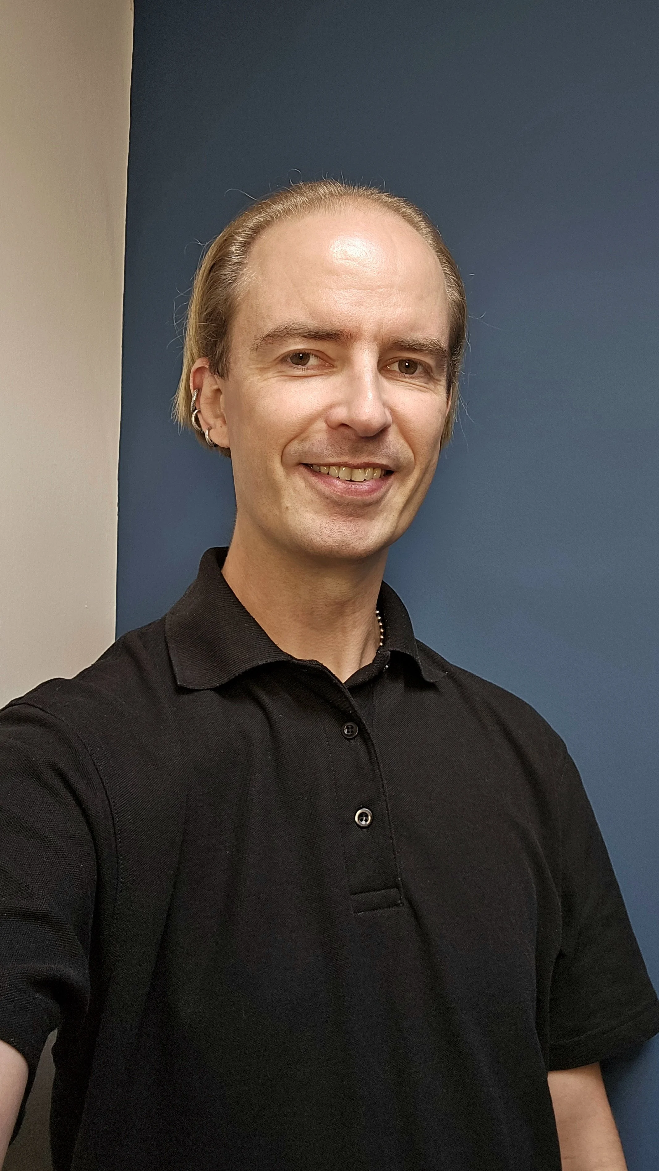A smiling man with light skin, wearing a black polo shirt and earrings, standing against a two-tone blue and beige background.