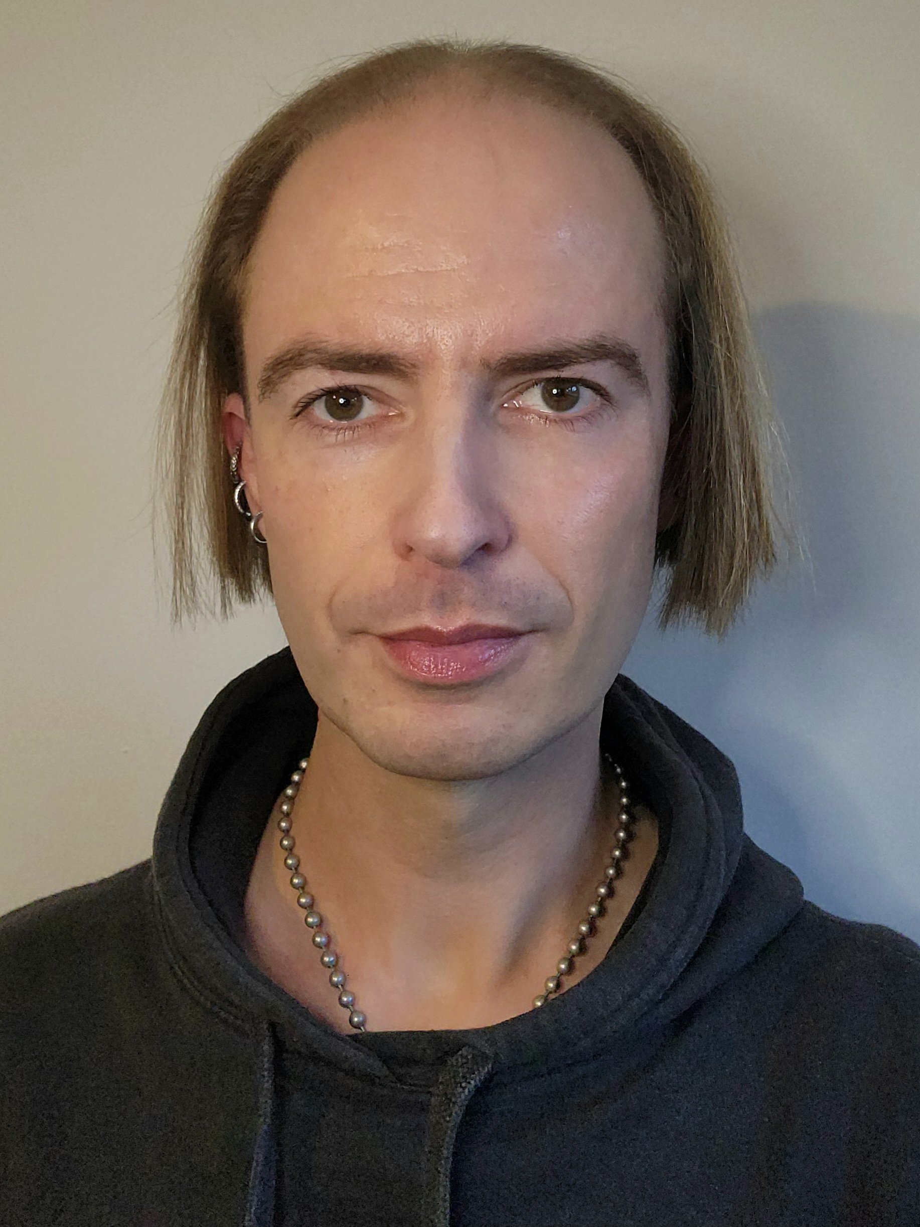 A person with shoulder-length brown hair, wearing earrings, a pearl necklace, and a black hoodie, looking directly at the camera with a neutral expression, standing against a plain, light-colored wall.