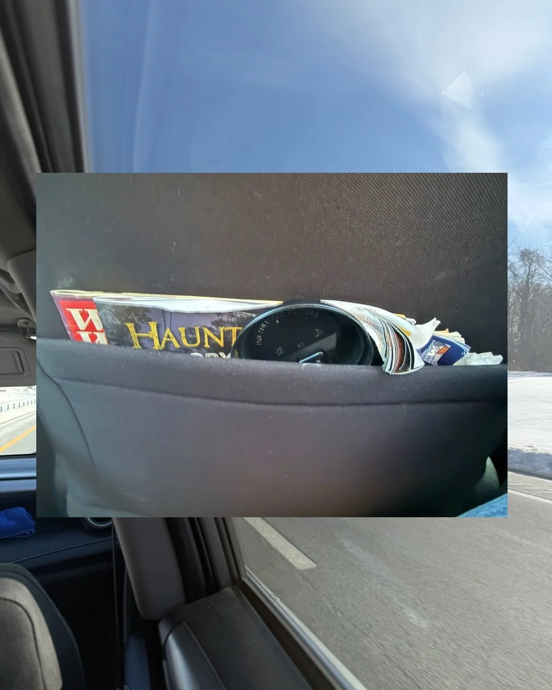 View of a car interior with a back seat pocket containing magazines, a black watch, and some papers with a snowy landscape outside the window.