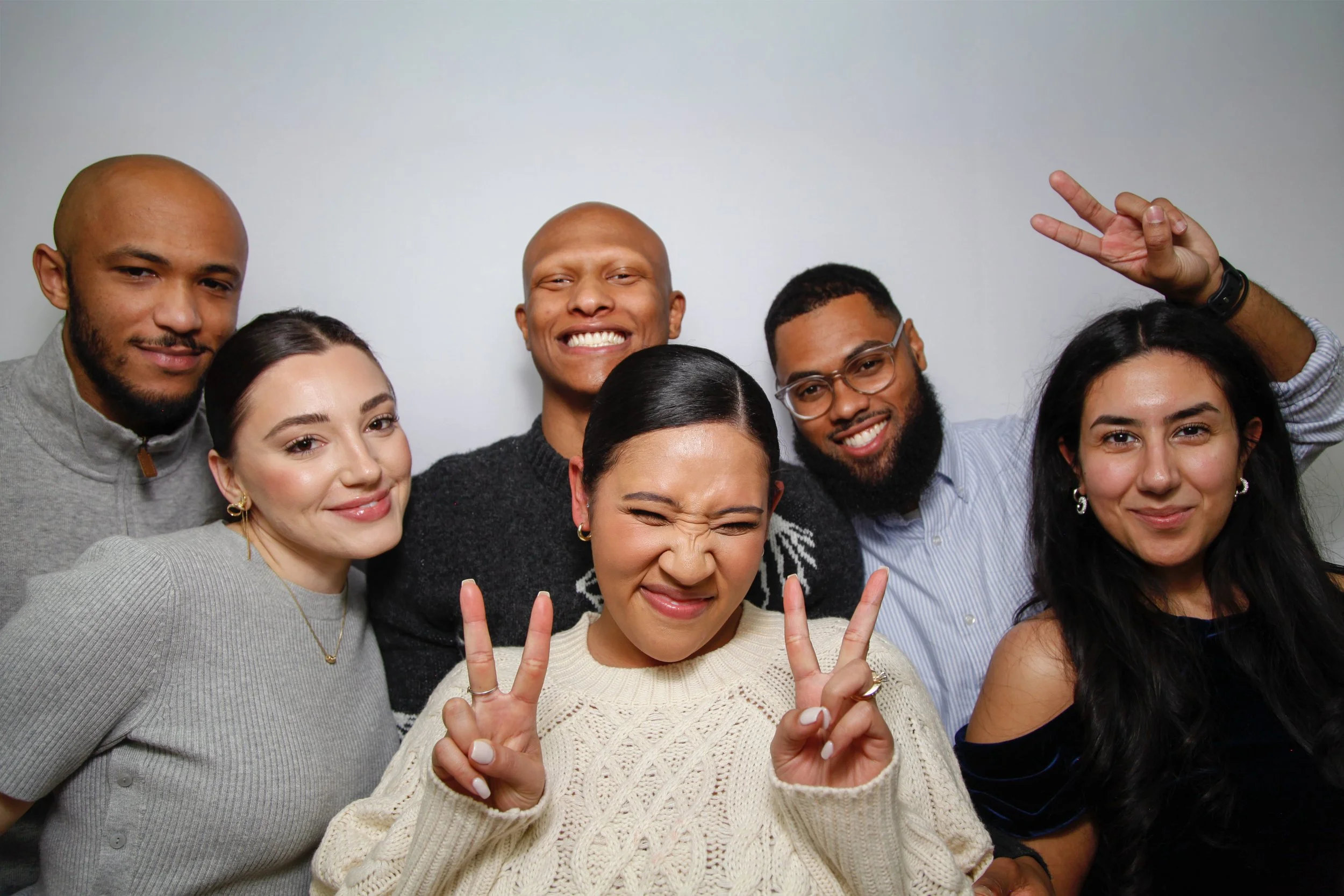 Group of seven diverse young adults smiling and making peace signs, standing close together against a plain, light-colored background.