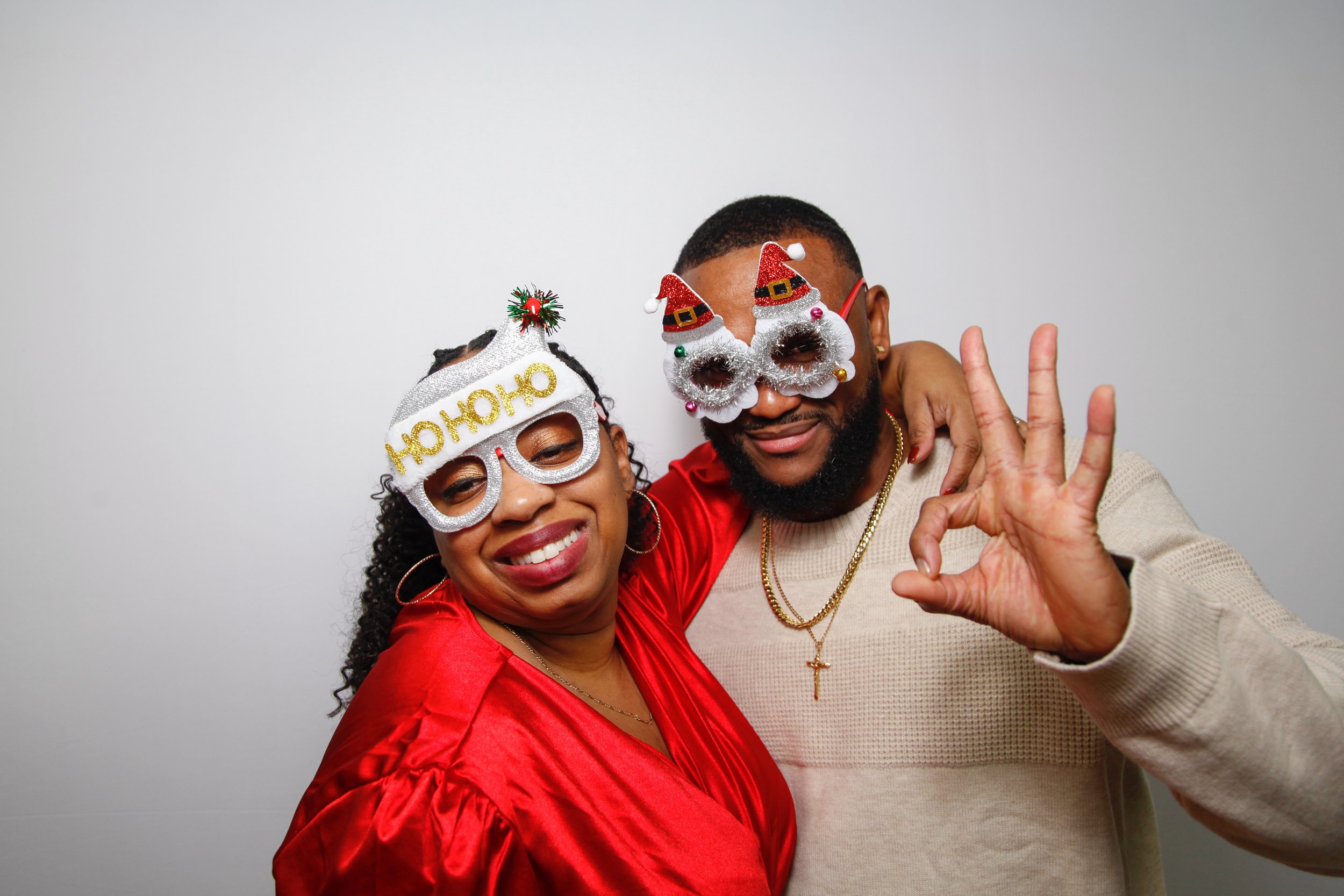 A smiling woman and man dressed festively for Christmas, wearing humorous holiday glasses, with the woman dressed in a red top and the man in a beige sweater with gold jewelry, posing together against a plain white background.