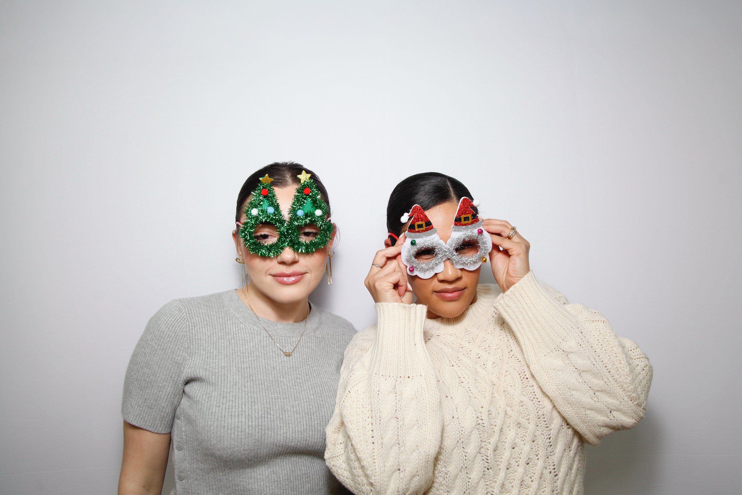 Two women wearing festive Christmas themed masks, one with green tinsel and the other with Santa and reindeer decorations, posing against a plain white background.