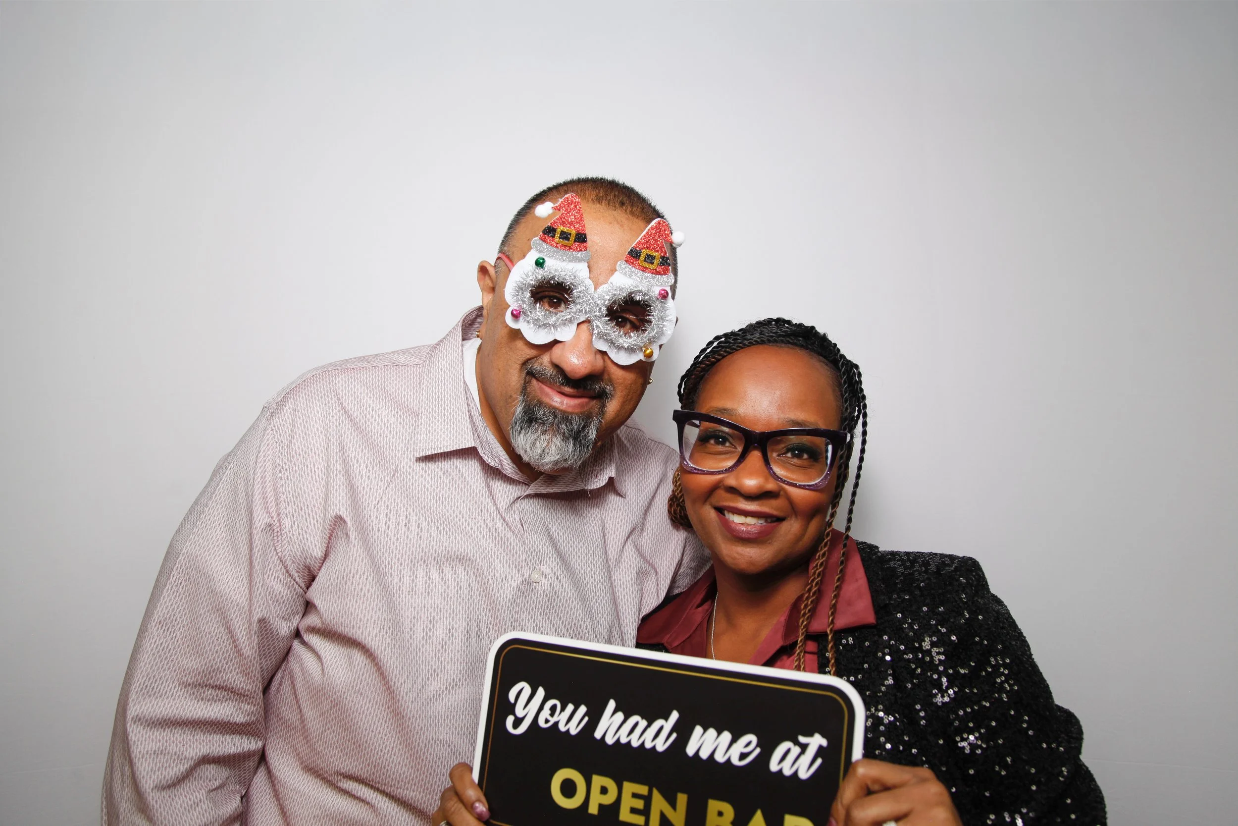 A man and woman smiling for a photo, the man wearing festive holiday glasses with Santa hats and the woman holding a sign that reads 'You had me at open bar'.