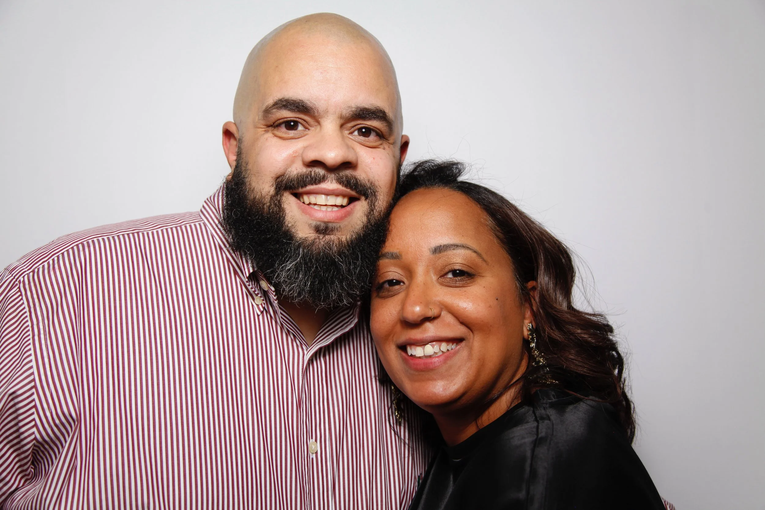 A smiling man with a bald head, beard, and striped shirt, standing next to a woman with dark hair, earrings, and a black top, against a plain background.