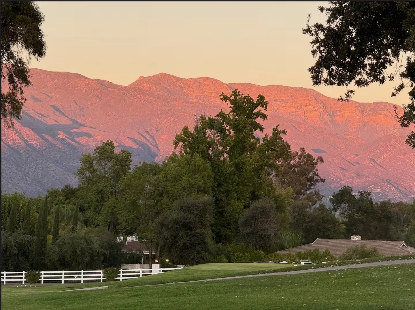 A scenic view of a suburban neighborhood with green lawns, trees, and houses in the foreground, and a mountain range illuminated by pinkish-orange sunlight in the background during sunset.