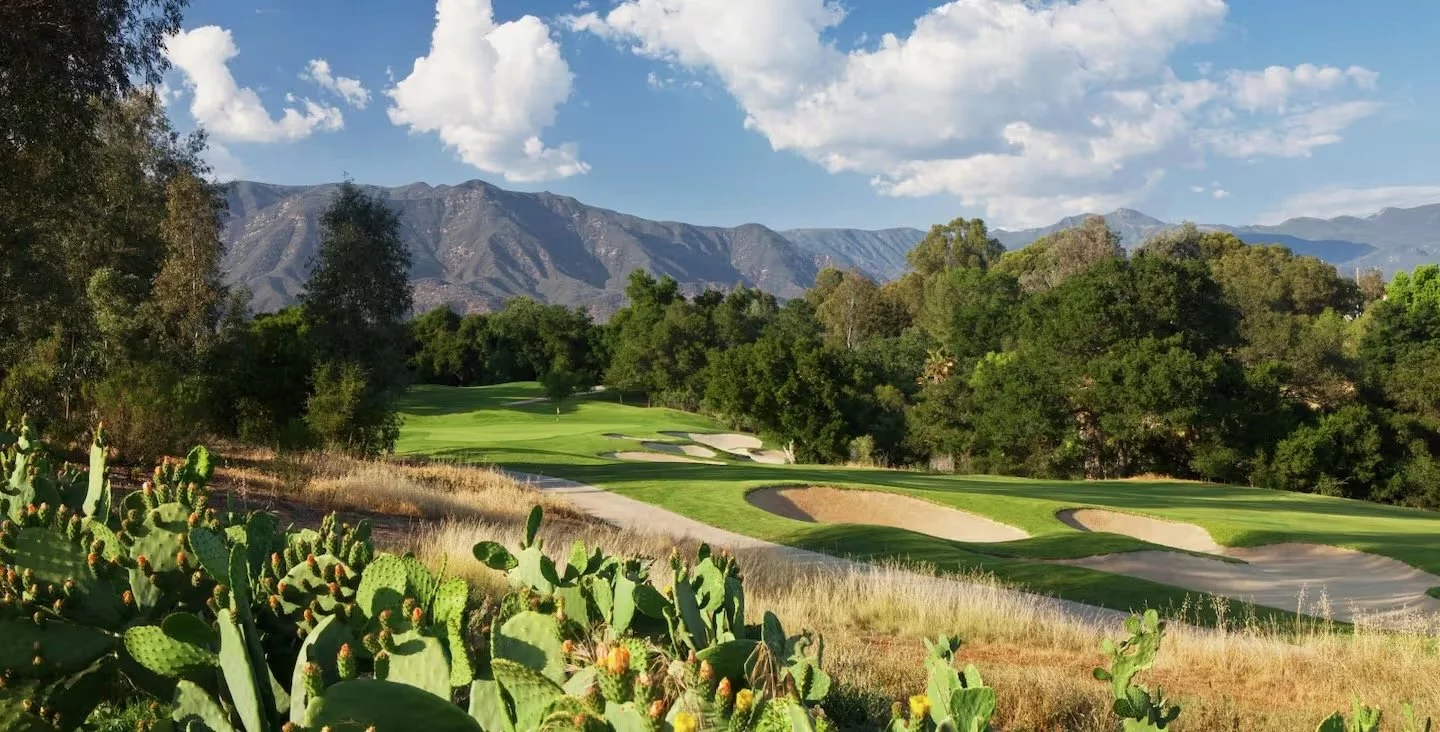 A golf course with sand traps, surrounded by trees and mountains in the background, under a blue sky with scattered clouds.