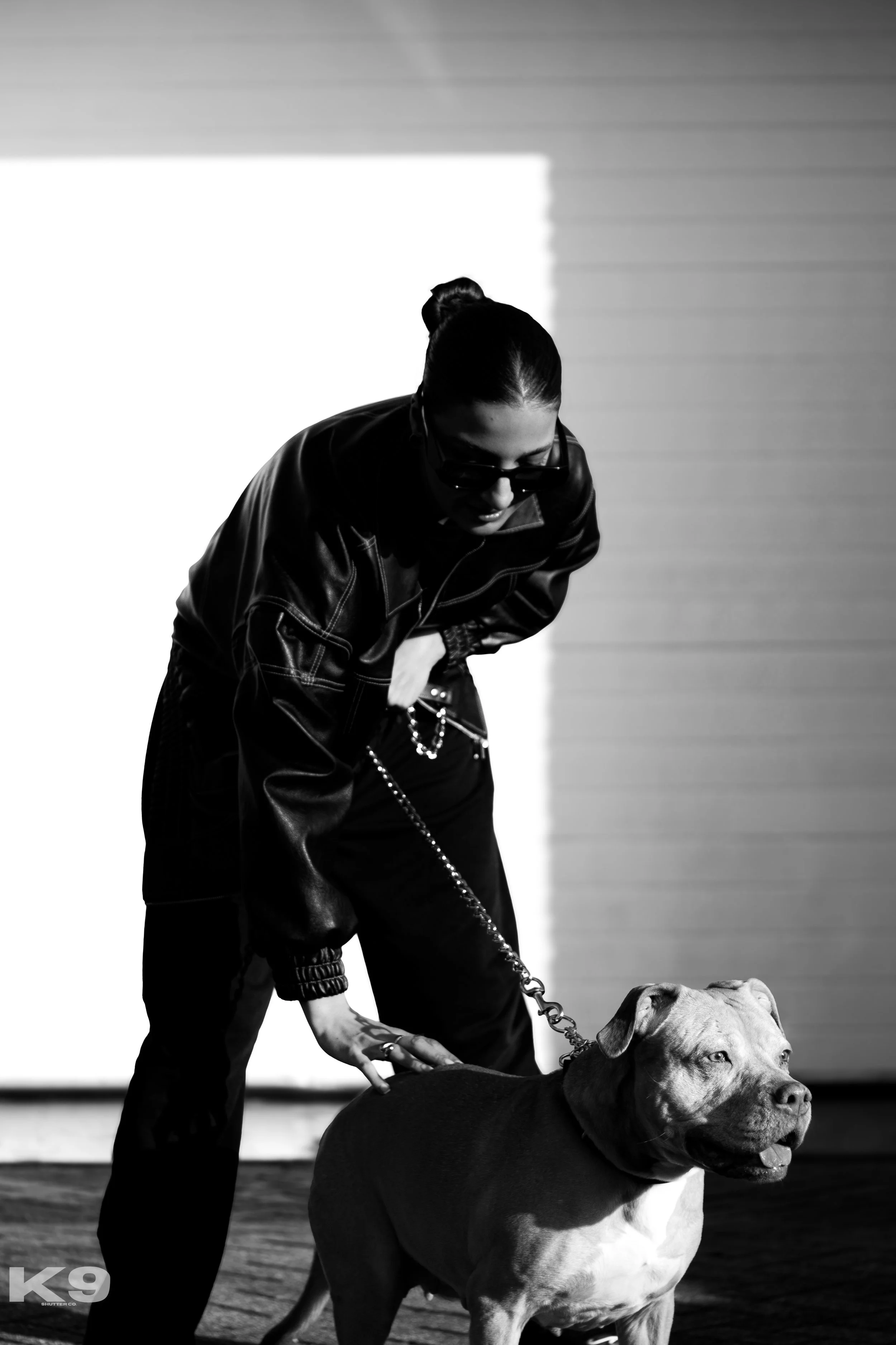 A woman wearing sunglasses and a leather jacket holding a chain leash attached to a dog, standing on a wooden floor in front of a plain background.