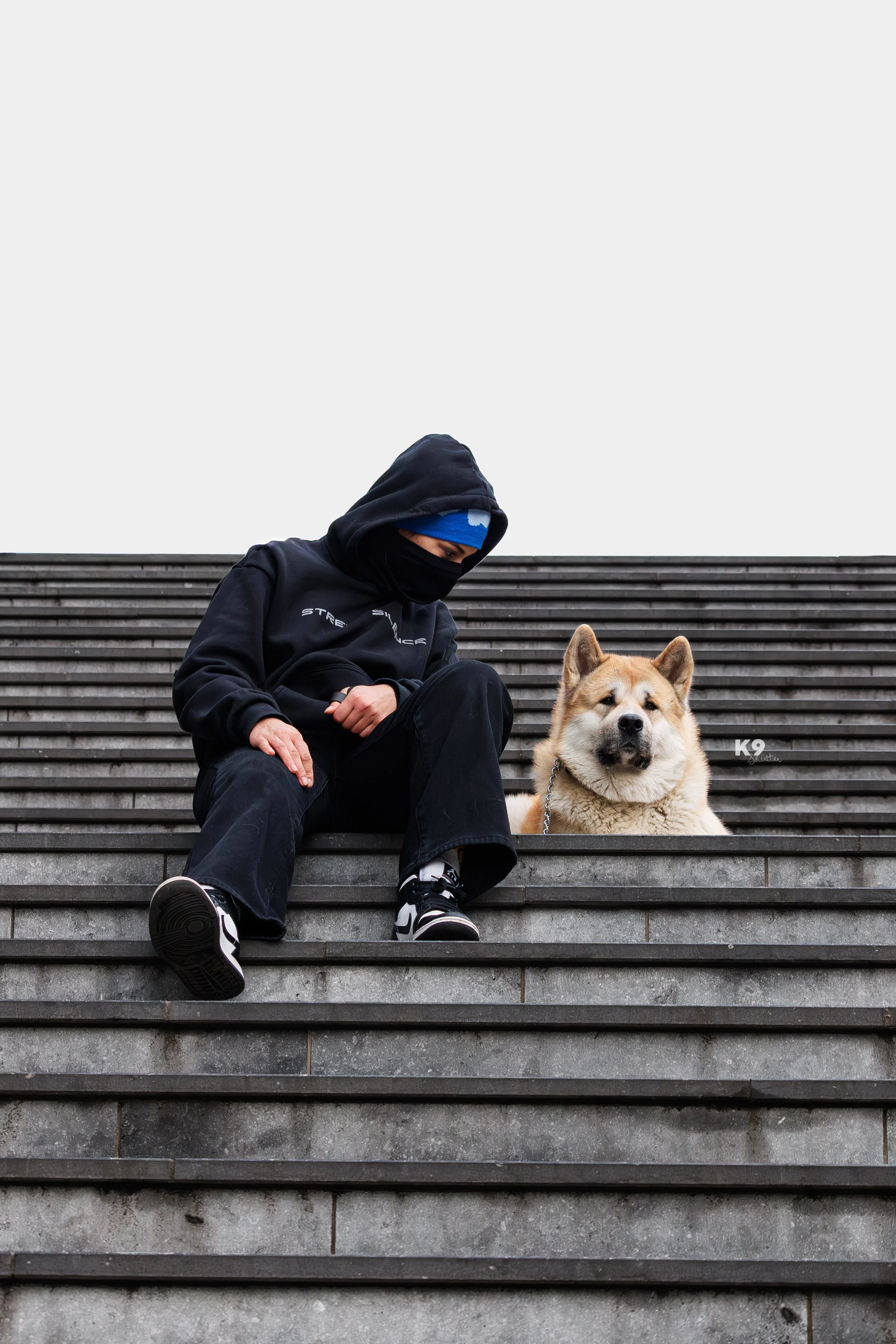 Person dressed in black hoodie, pants, and mask sitting on outdoor stairs next to a large light-colored dog, possibly a husky or Akita, with the sky in the background.