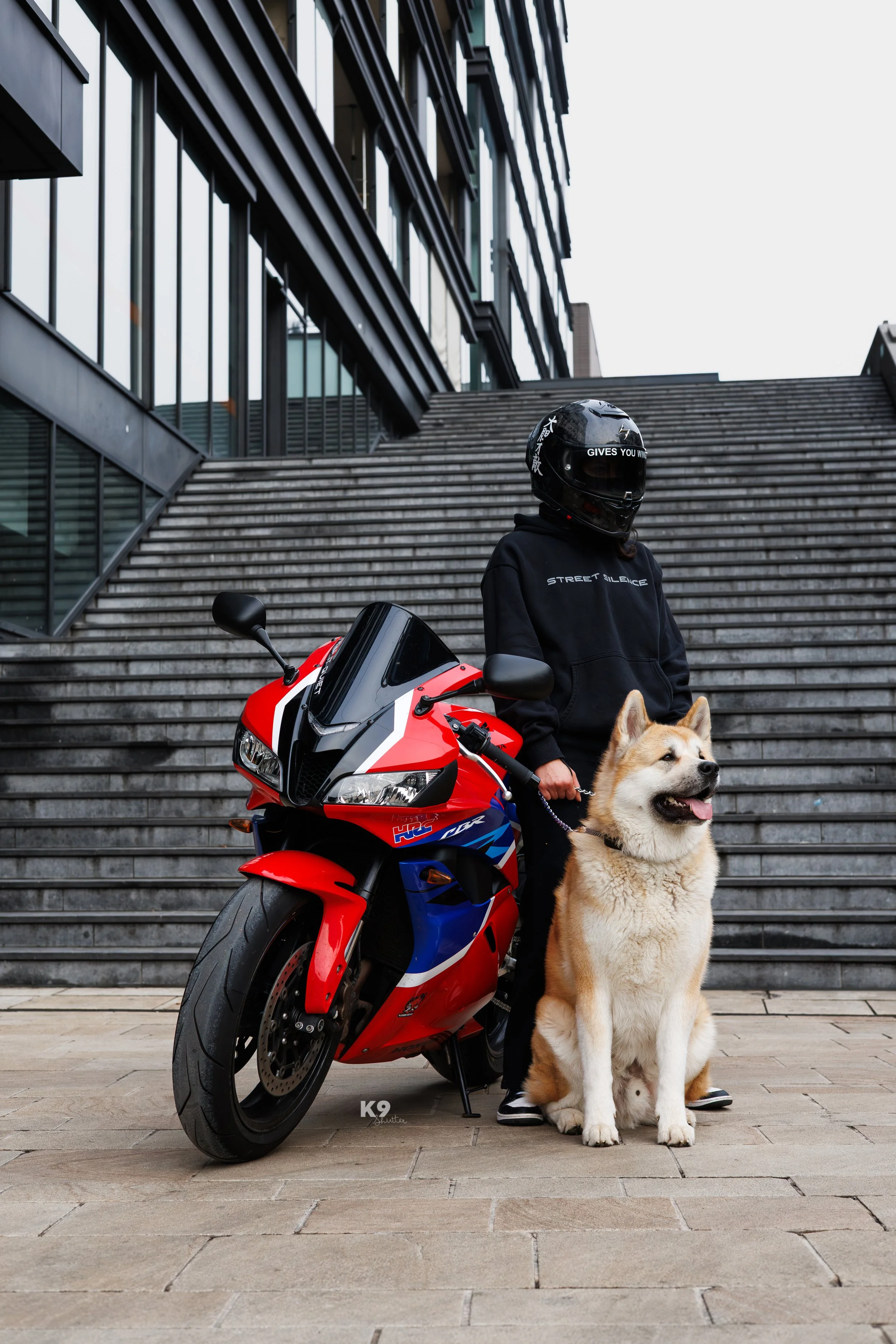 Person in black hoodie and helmet standing next to red, white, and blue sports motorcycle, holding a leash attached to large tan and white dog sitting on a paved surface in front of stairs and modern building.