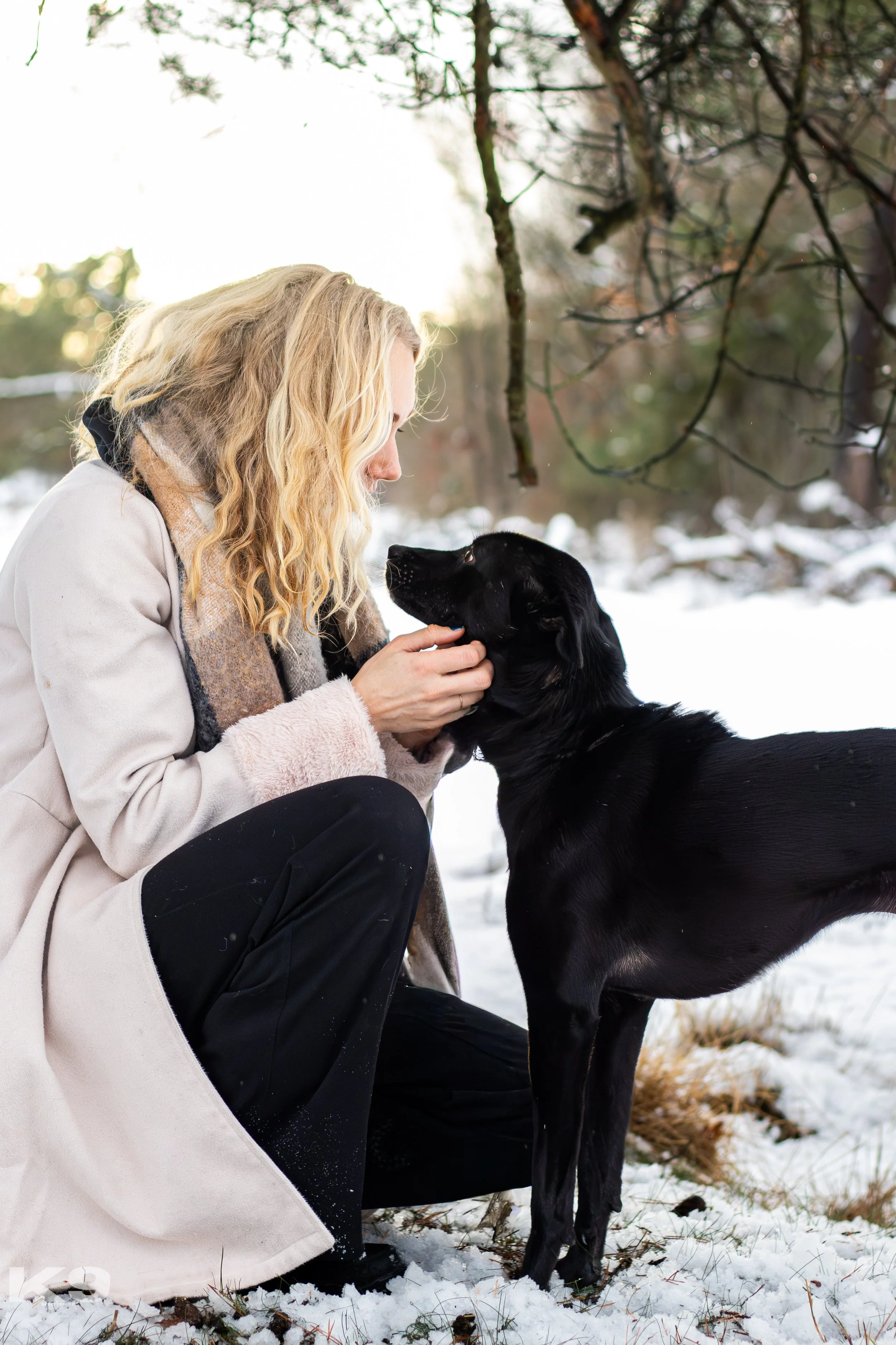 A woman with blonde hair kneeling in the snow and holding a black dog, looking into its eyes outdoors in a winter setting.