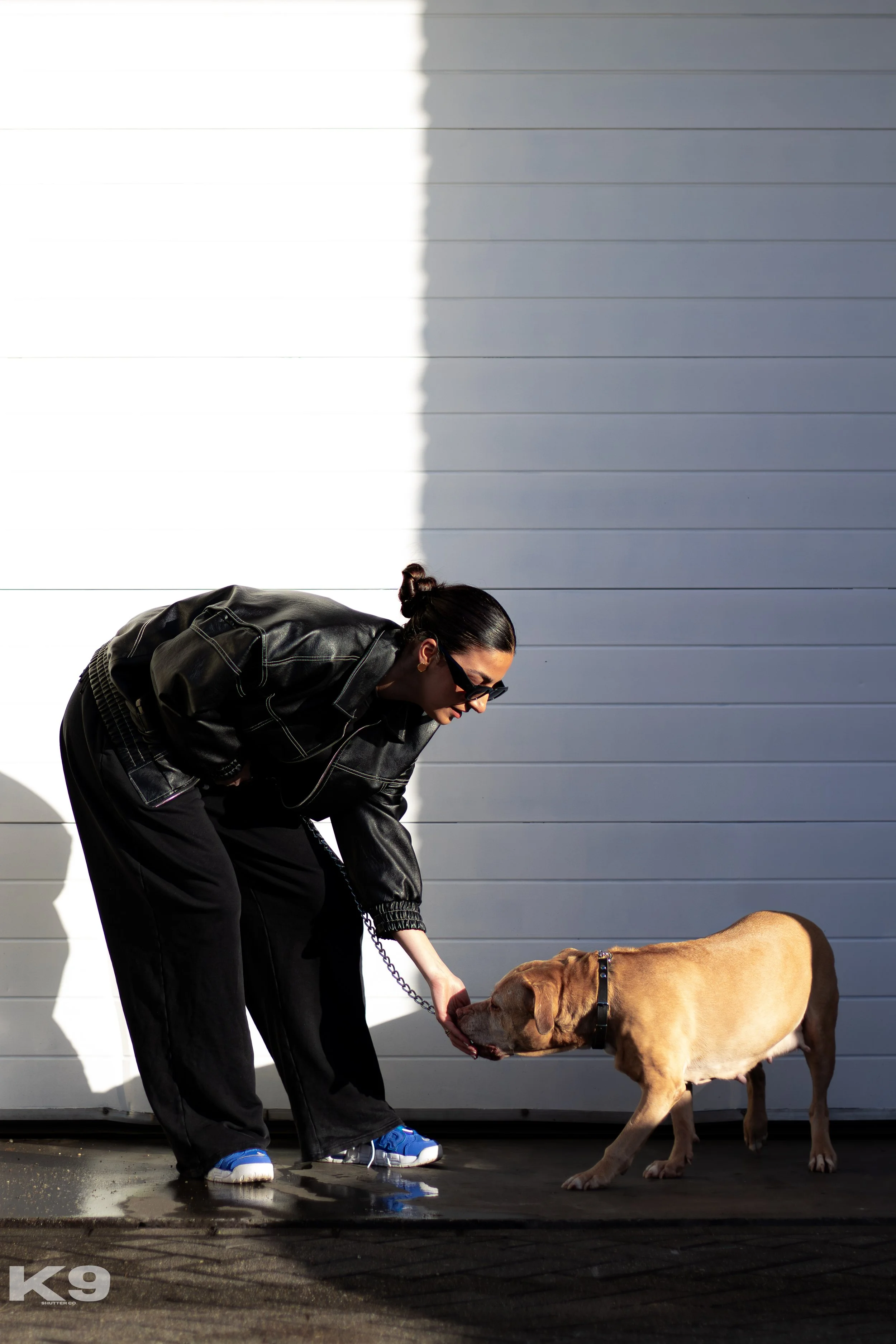 A woman in a black leather jacket and sunglasses crouches down to pet a large brown dog with a black collar against a grey garage door.