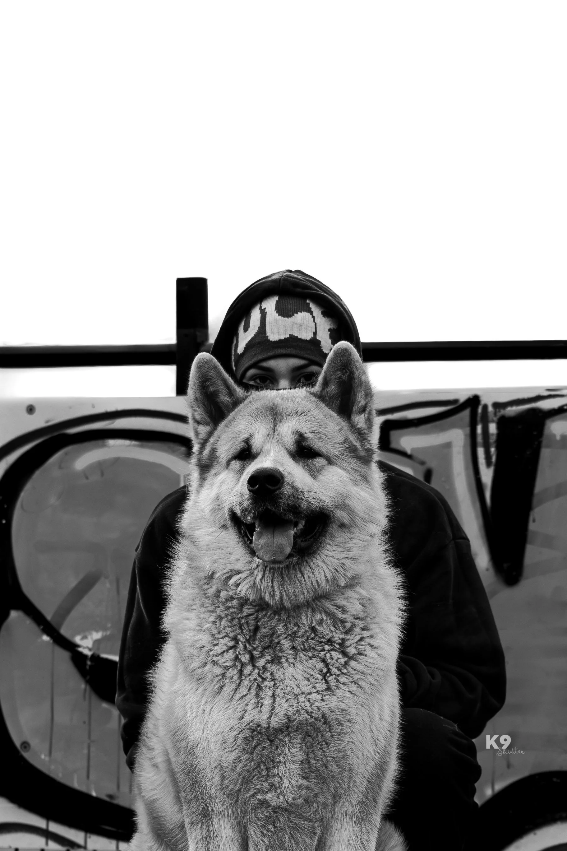 A person wearing a camouflage-patterned hat and dark clothing is partially hidden behind a large, fluffy dog with a happy expression, in front of a graffiti-covered wall.
