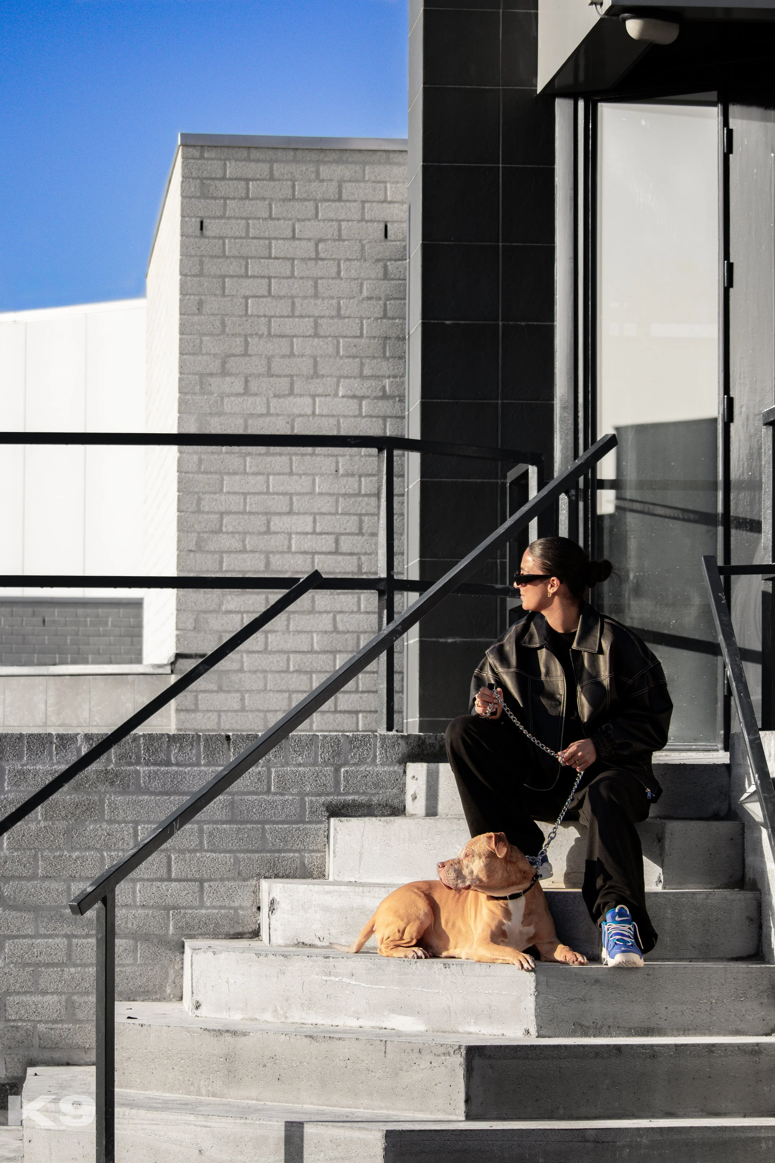 A woman with dark hair tied in a bun is sitting on concrete steps outside a building, holding a chain connected to a tan dog laying at her feet. She is wearing sunglasses, a black jacket, and sneakers, with a modern building and a clear blue sky in t