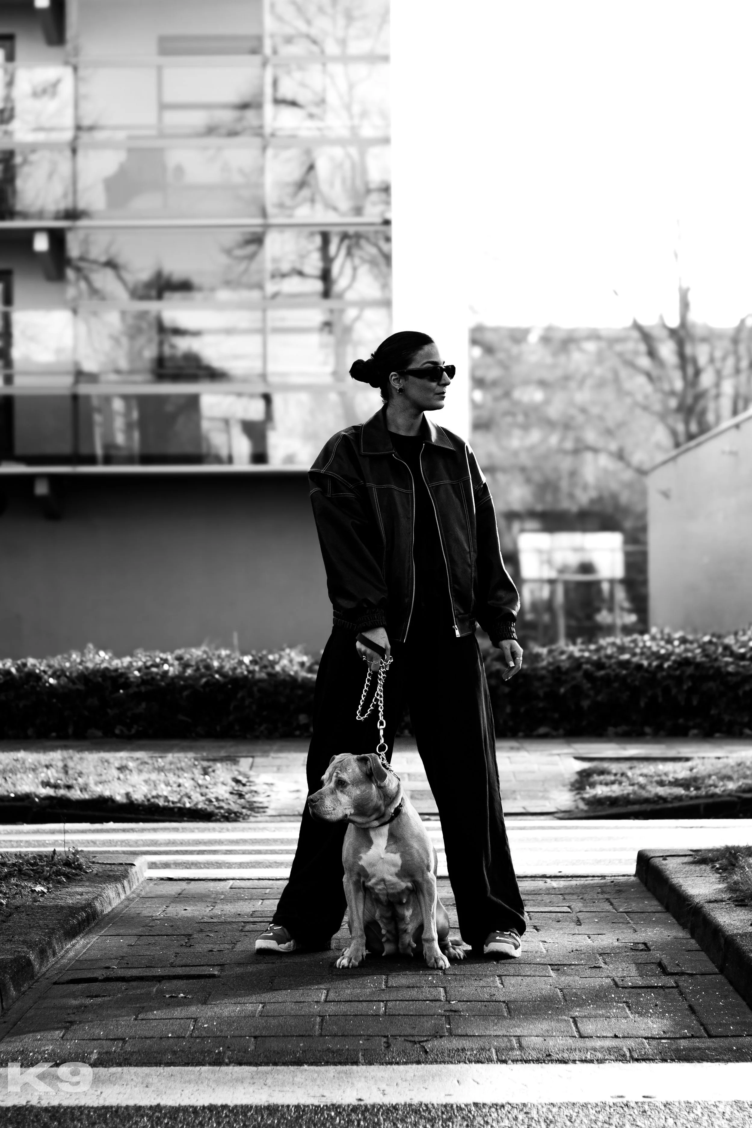 A woman wearing sunglasses and a leather jacket stands on a crosswalk with her dog on a leash in an urban area. The photo is in black and white.