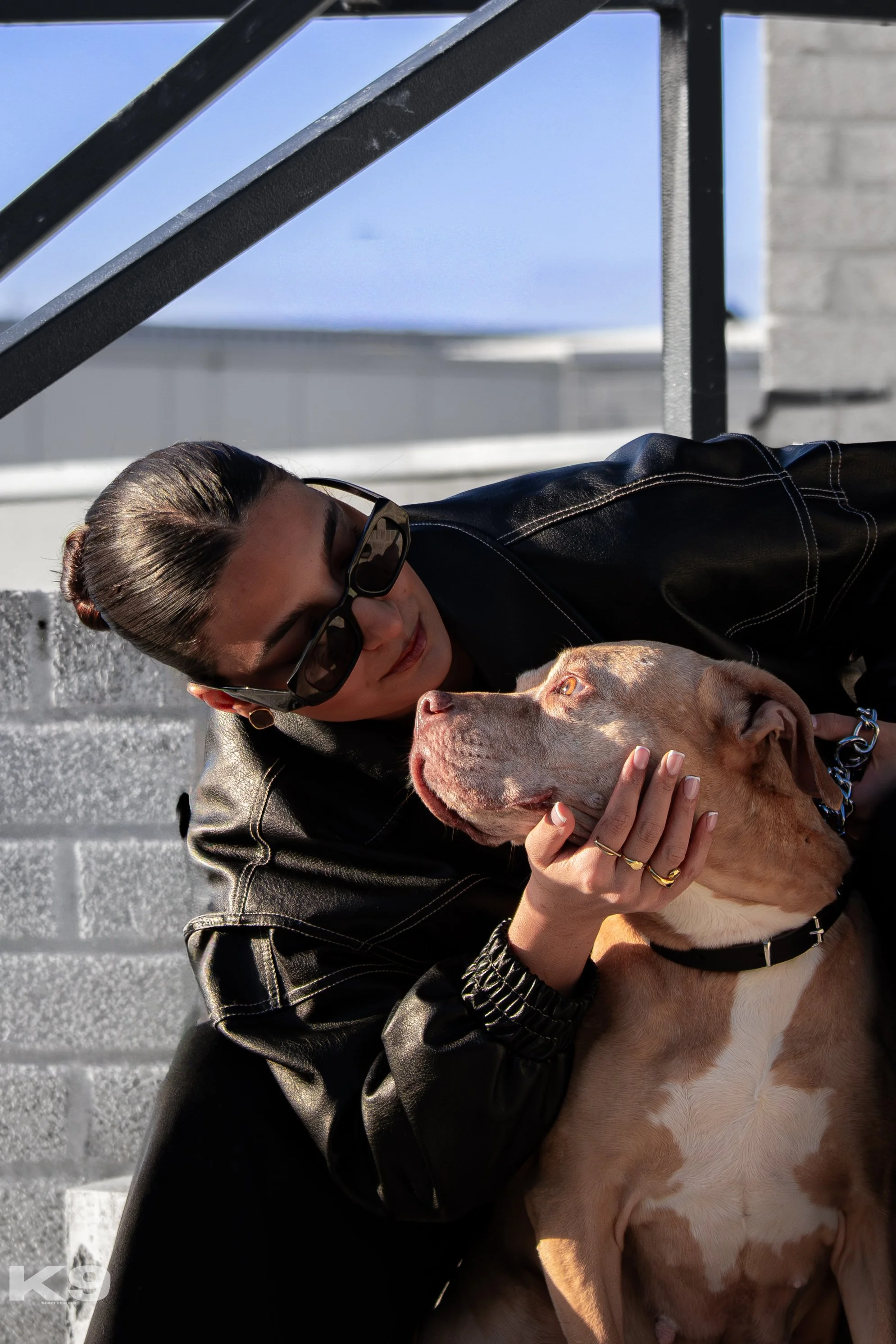 A woman wearing sunglasses and a black leather jacket is petting a large brown dog with amber eyes, sitting outside on a sunny day near a brick wall and metal railing.