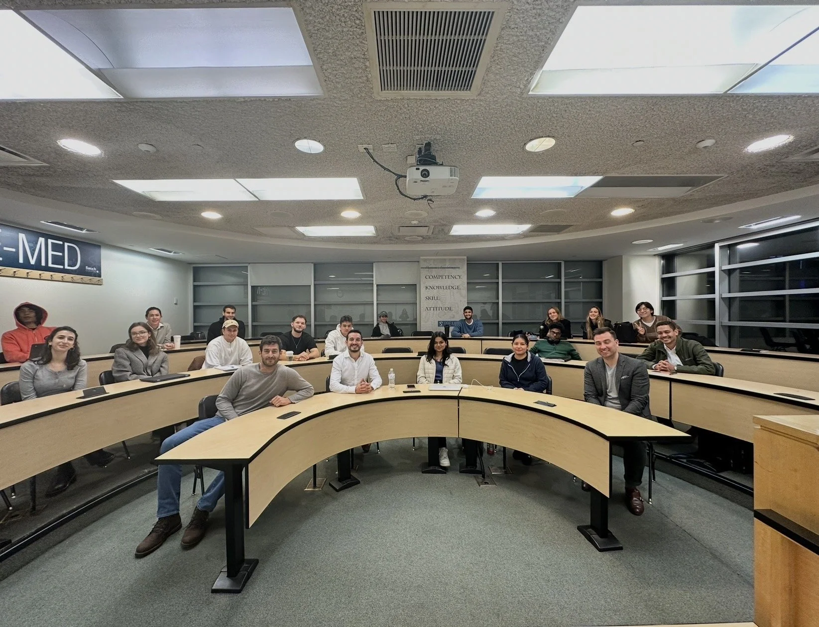 A diverse group of students seated in a lecture hall, facing the camera, with a large digital screen on the left and a whiteboard on the back wall. The room has multiple rows of curved desks and overhead lights.