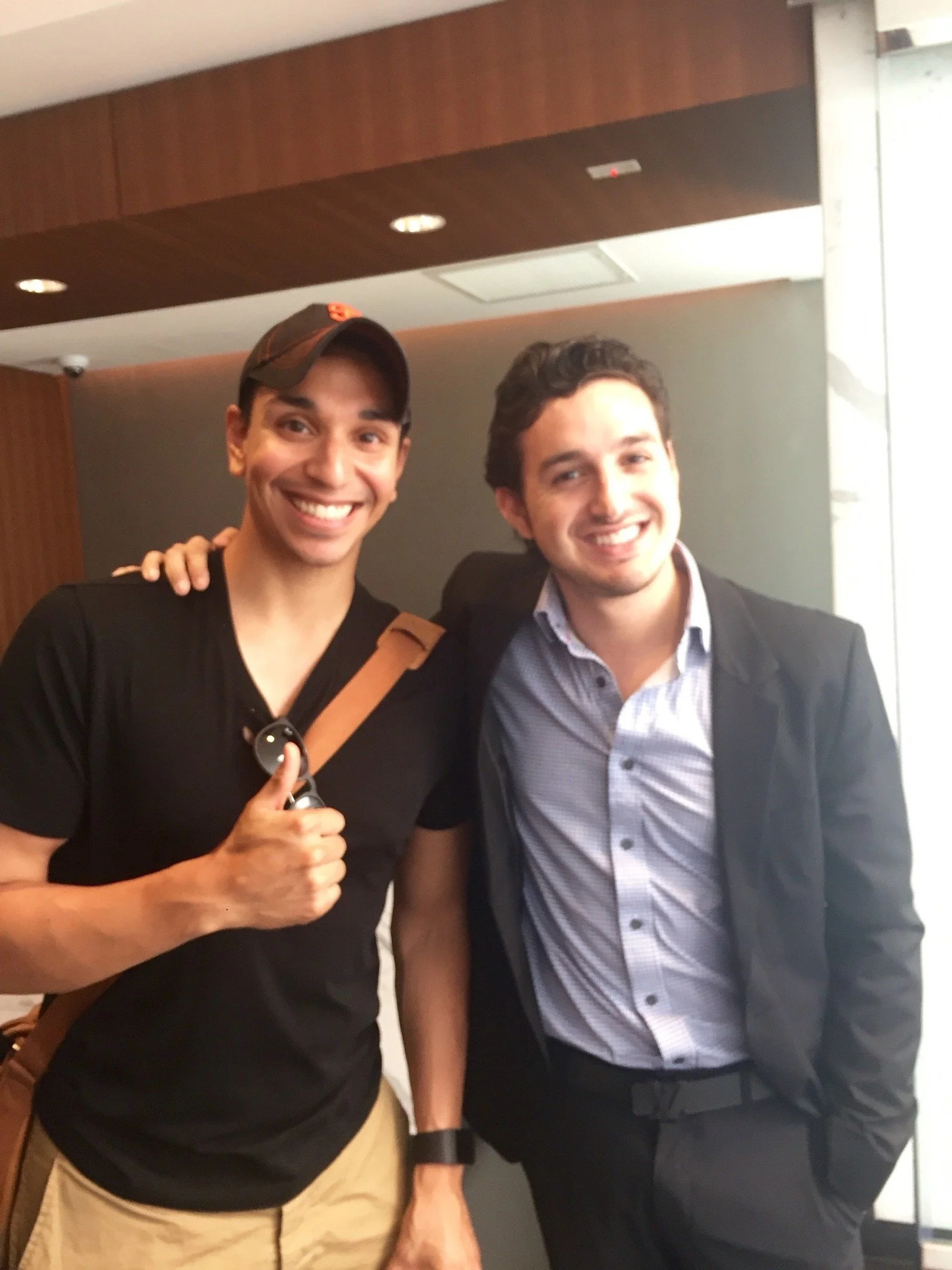 Two young men smiling and posing for a photo indoors, one giving a thumbs-up, with the other resting his arm on his friend's shoulder.