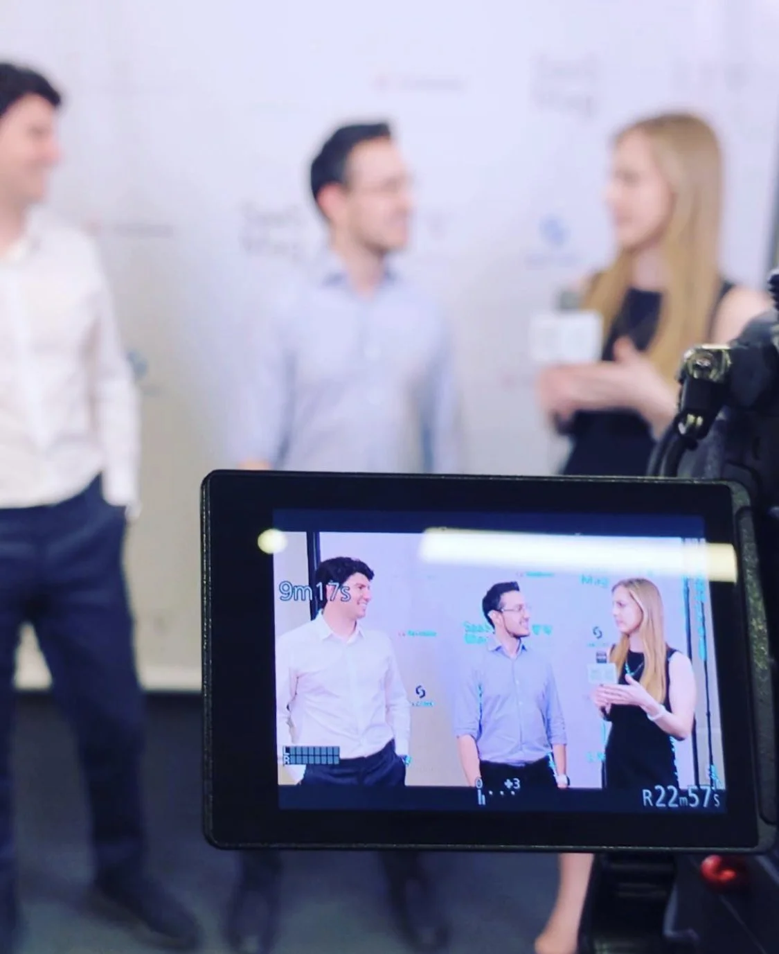 A camera's screen showing three people being filmed against a white background, with a woman holding papers talking to two men, all dressed in business casual attire.