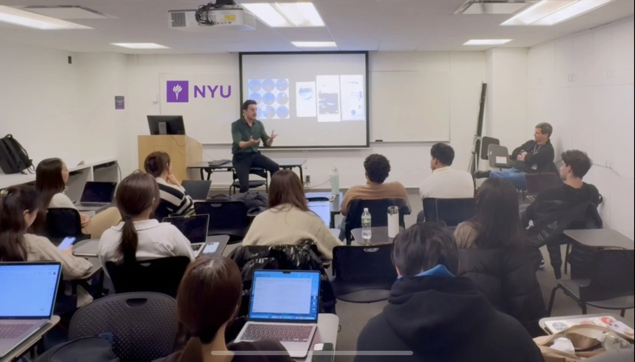 A classroom at NYU with a teacher standing in front of students, presenting slides on a screen. Students are seated at desks with laptops and water bottles, listening attentively.