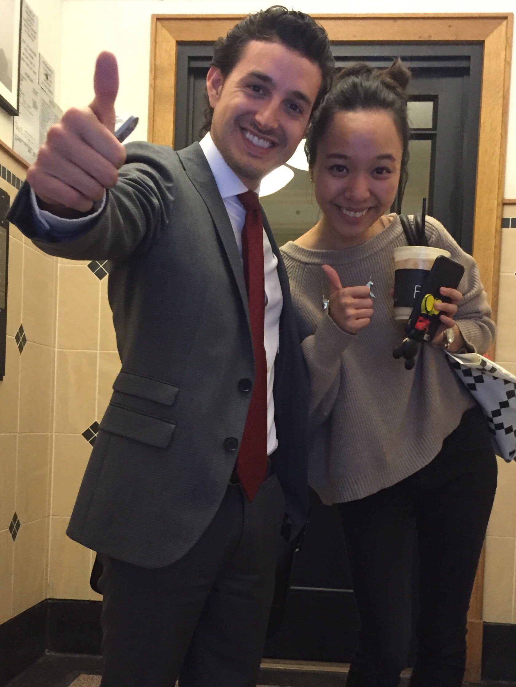 A man in a gray suit and red tie giving a thumbs up next to a woman in a beige sweater holding a drink and also giving a thumbs up, both smiling indoors.