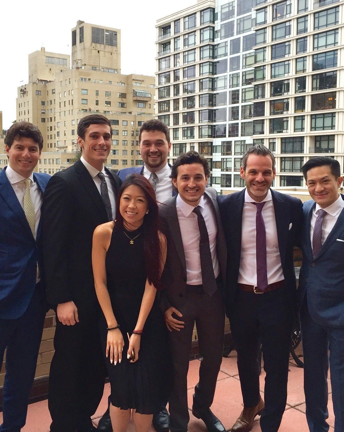 Group of eight young professionals in formal attire standing on a balcony with city buildings in the background.