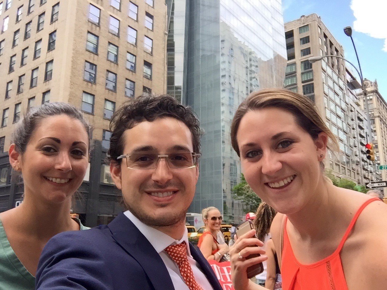 Three people smiling at the camera on a city street with tall buildings in the background. One woman on the left, one man in the center wearing glasses and a suit, and another woman on the right in an orange top.