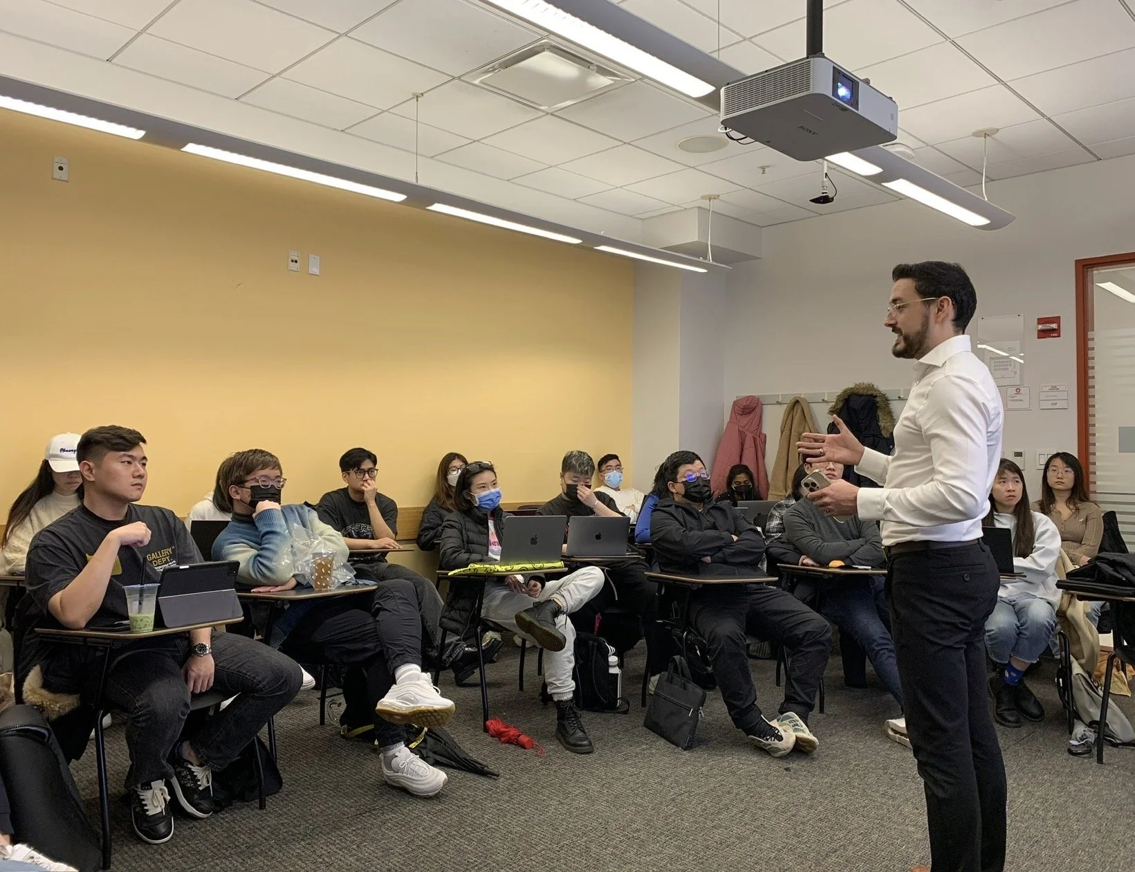 A classroom or lecture hall with students sitting at desks, some wearing face masks, listening to a male instructor who is standing and speaking while gesturing with his hands.