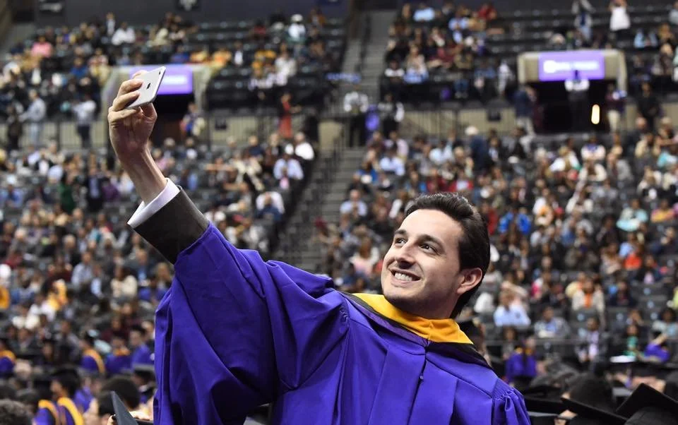 A graduate in a purple cap and gown taking a selfie at a graduation ceremony.