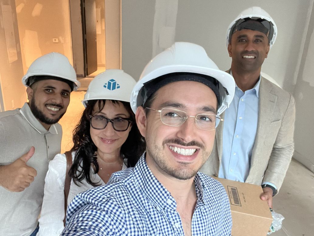 Group of four diverse construction professionals wearing white safety helmets, smiling, in an unfinished building.