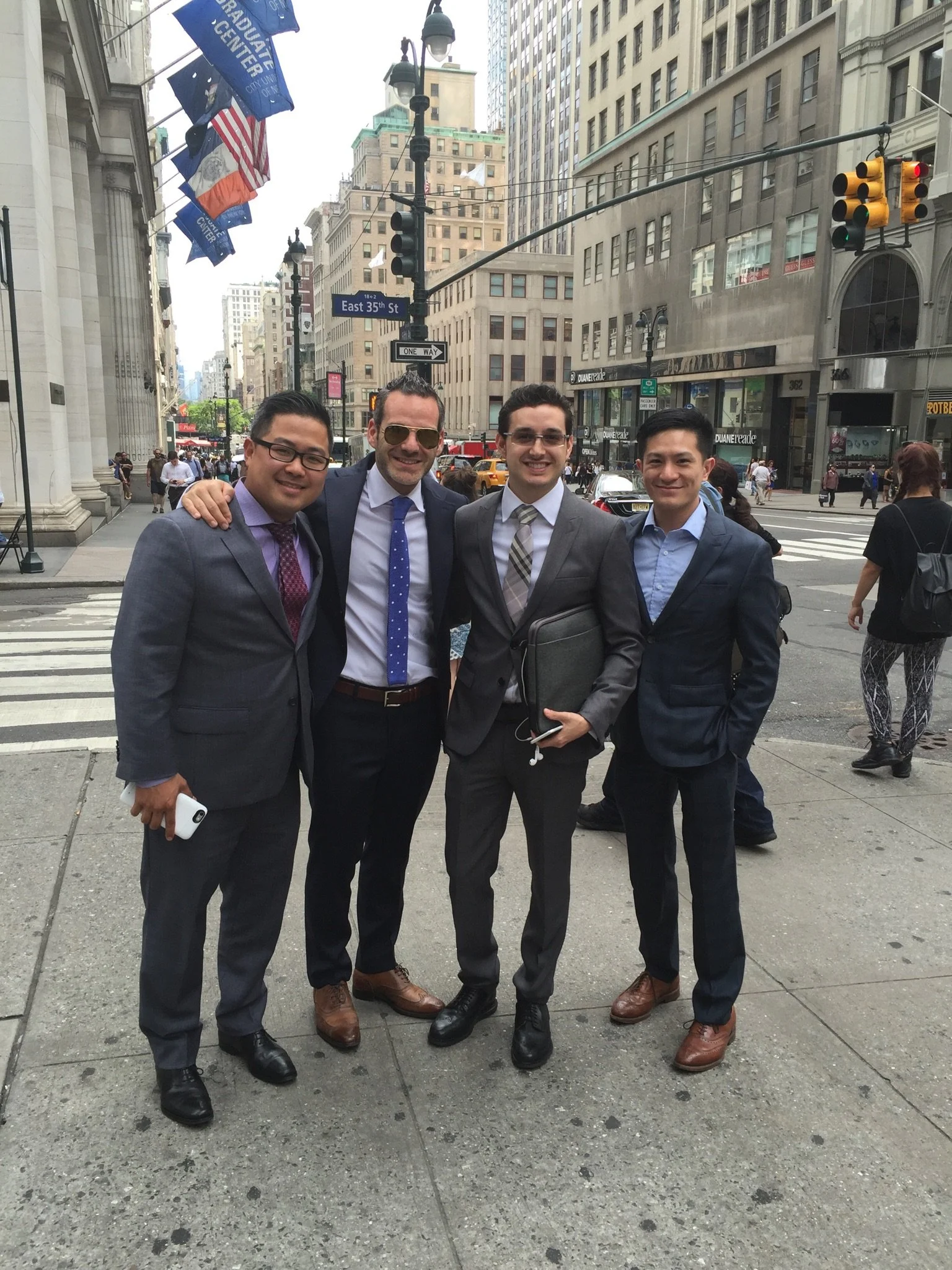Four men in suits standing on a city street corner, smiling for the camera, with tall buildings and traffic lights in the background.