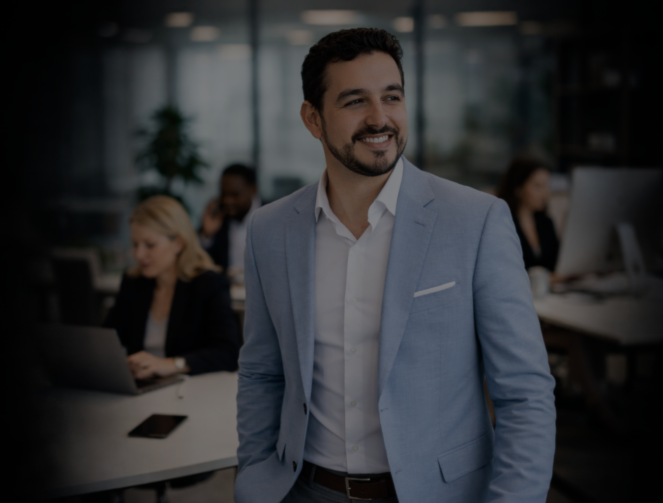 A man in a light blue suit smiling in a modern office with coworkers working on laptops in the background.