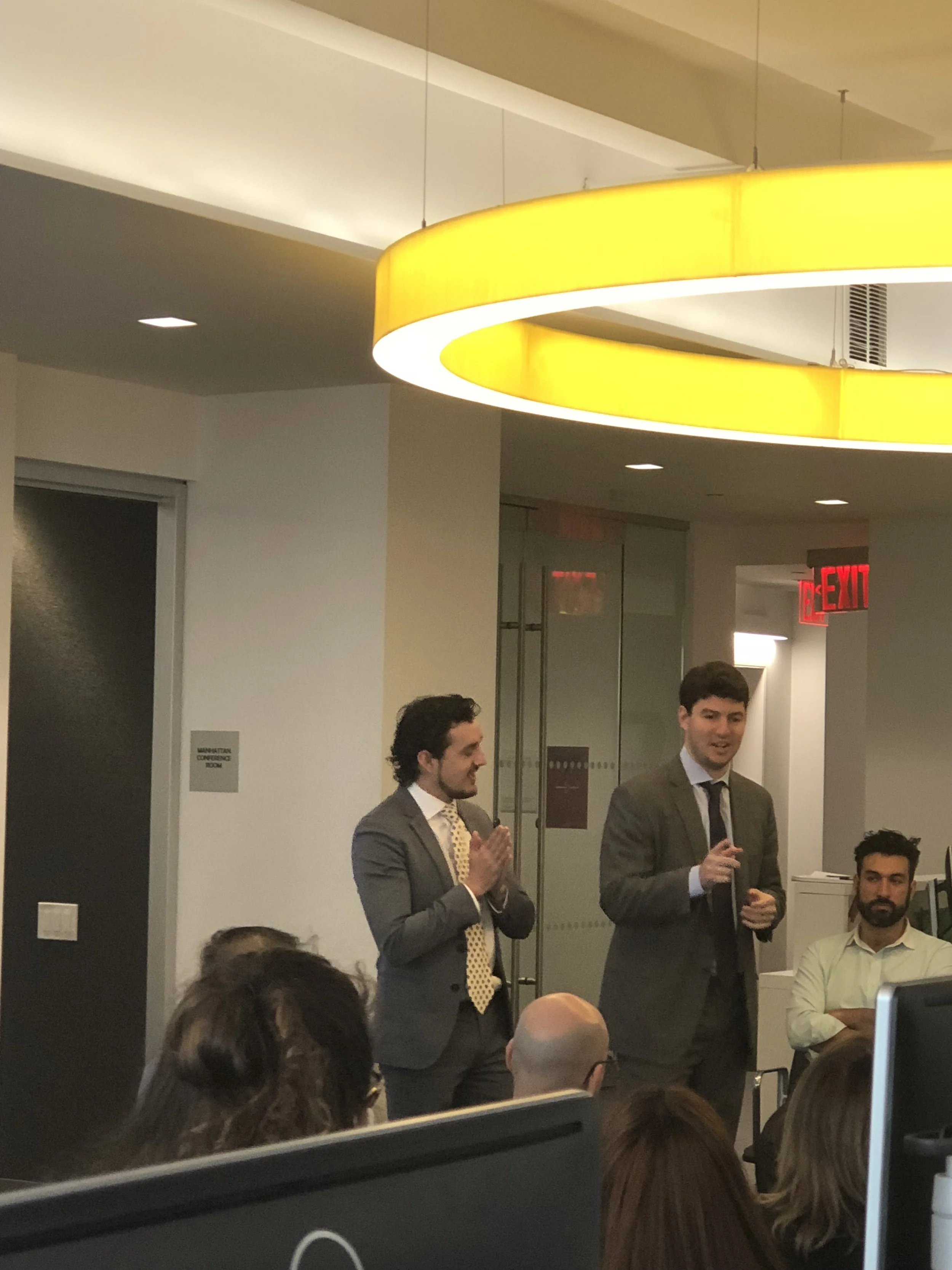 Two men in suits standing in front of an audience in an office conference room. One is speaking, the other is clapping, with many people seated watching them. There is a bright yellow circular ceiling light and an exit sign in the background.