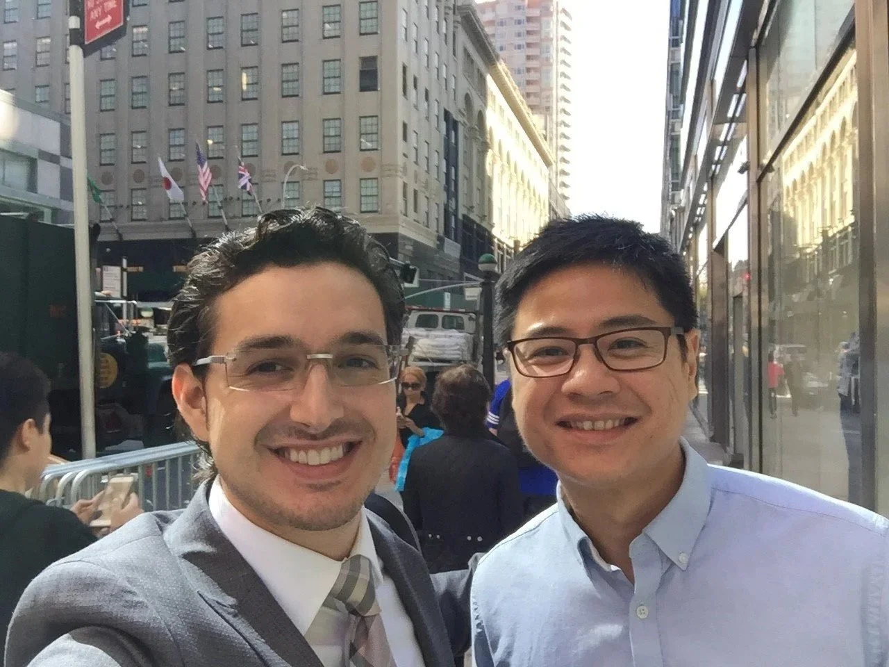 Two men in glasses smiling outdoors on a city street with tall buildings and pedestrians.