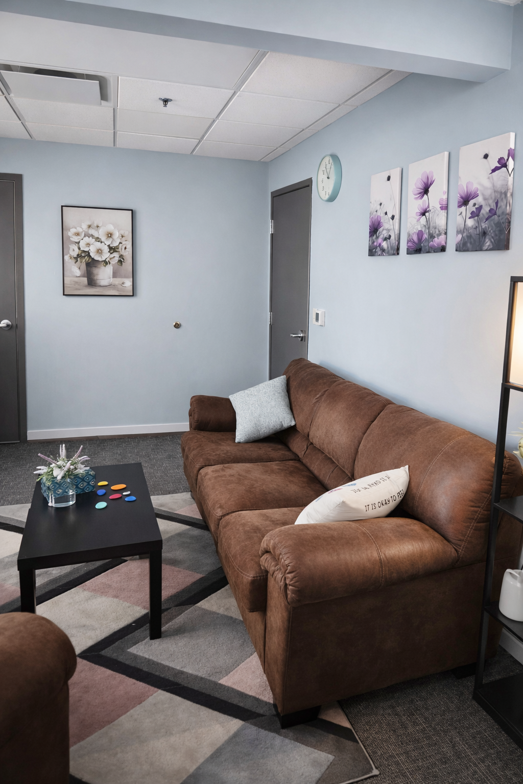 Living room with a brown sofa, a black coffee table with colorful buttons and a small flower arrangement, wall art with purple flowers, a blue clock, and a framed picture of white flowers on a light blue wall.