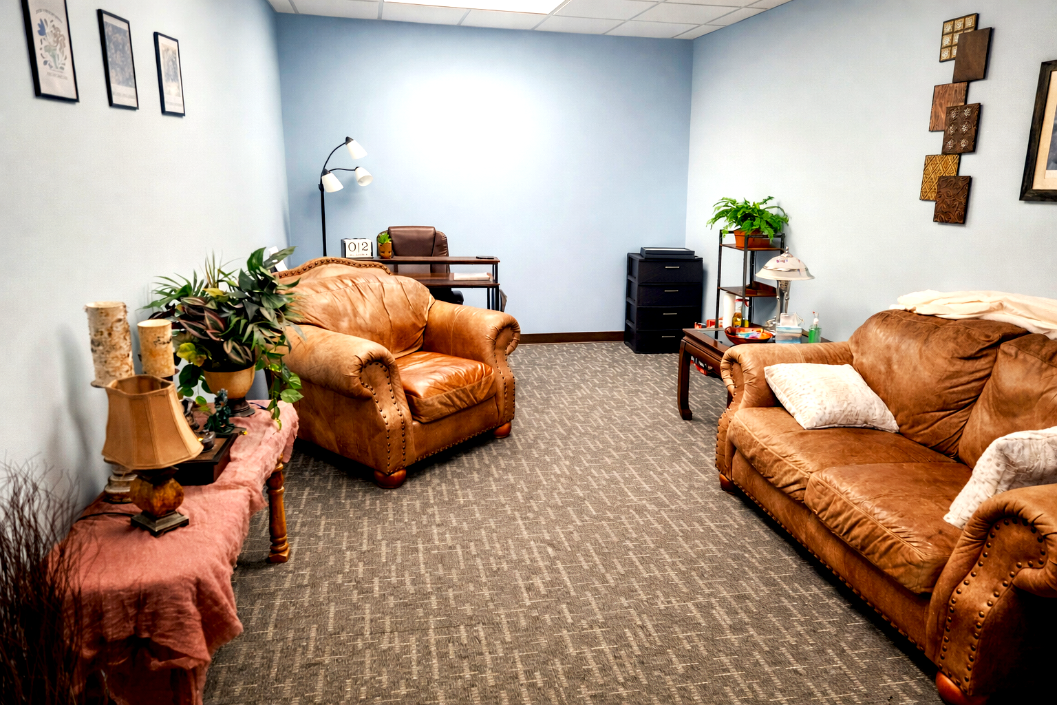 A cozy waiting room with light blue walls, featuring brown leather sofas, a side table with decorative items, a plant, a desk with a chair, and wall art.