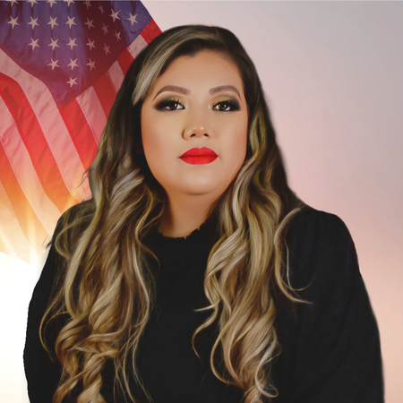 A woman with long wavy hair, red lipstick, and eye makeup, sitting in front of an American flag.