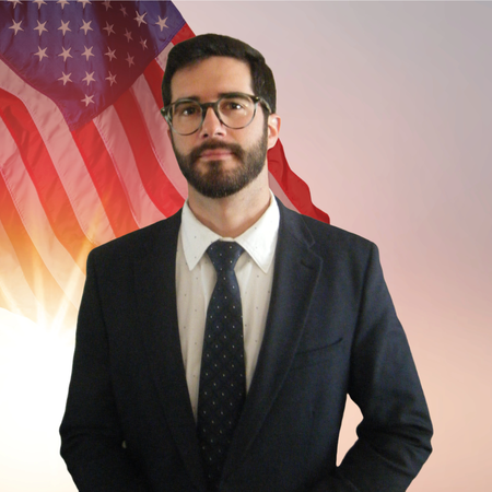 A man with glasses, dark hair and beard wearing a suit and tie standing in front of an American flag.