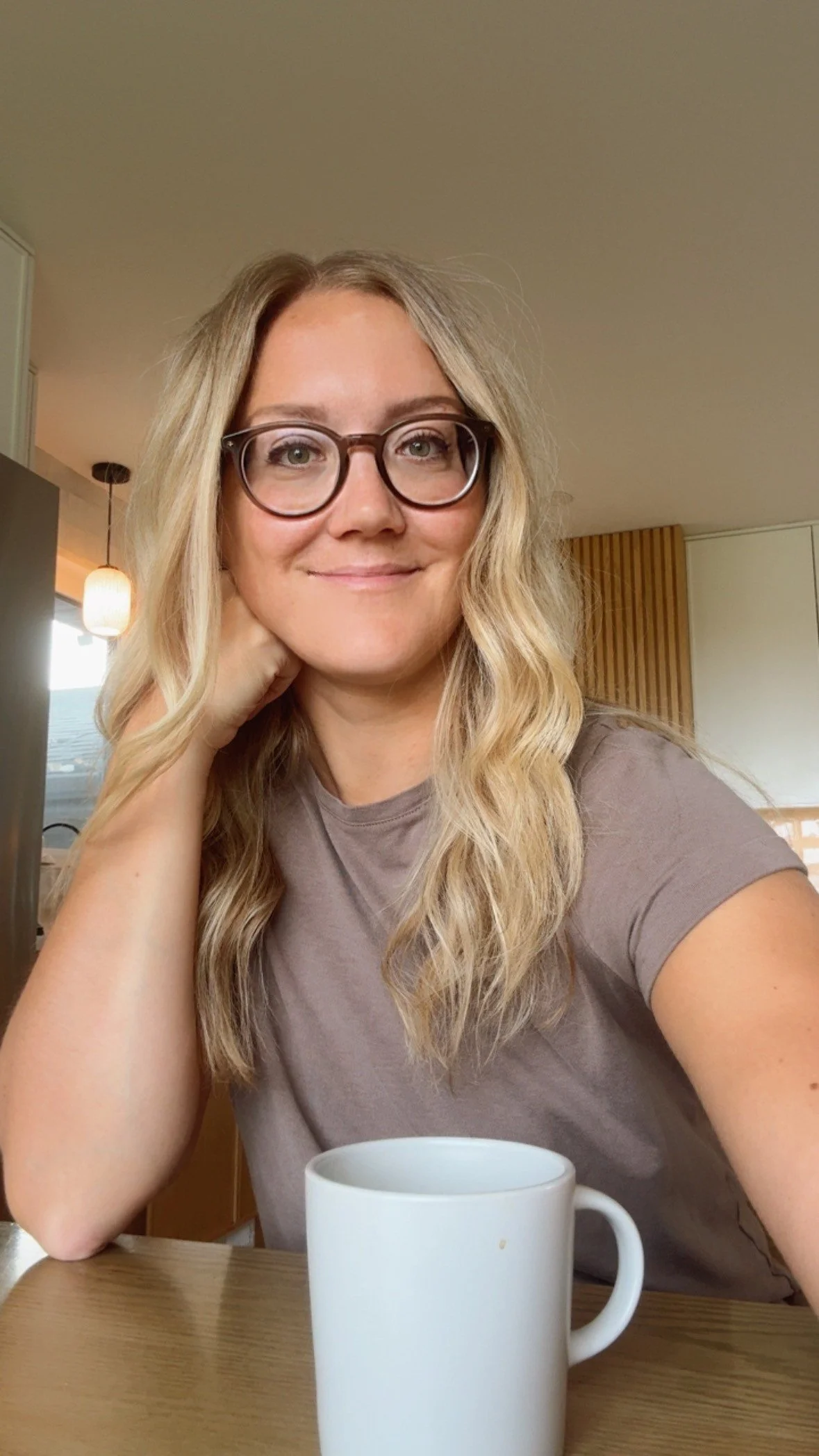 A woman with blonde wavy hair and glasses, smiling, sitting at a wooden table with a white mug in front of her, in a cozy indoor setting with warm lighting and a modern interior.
