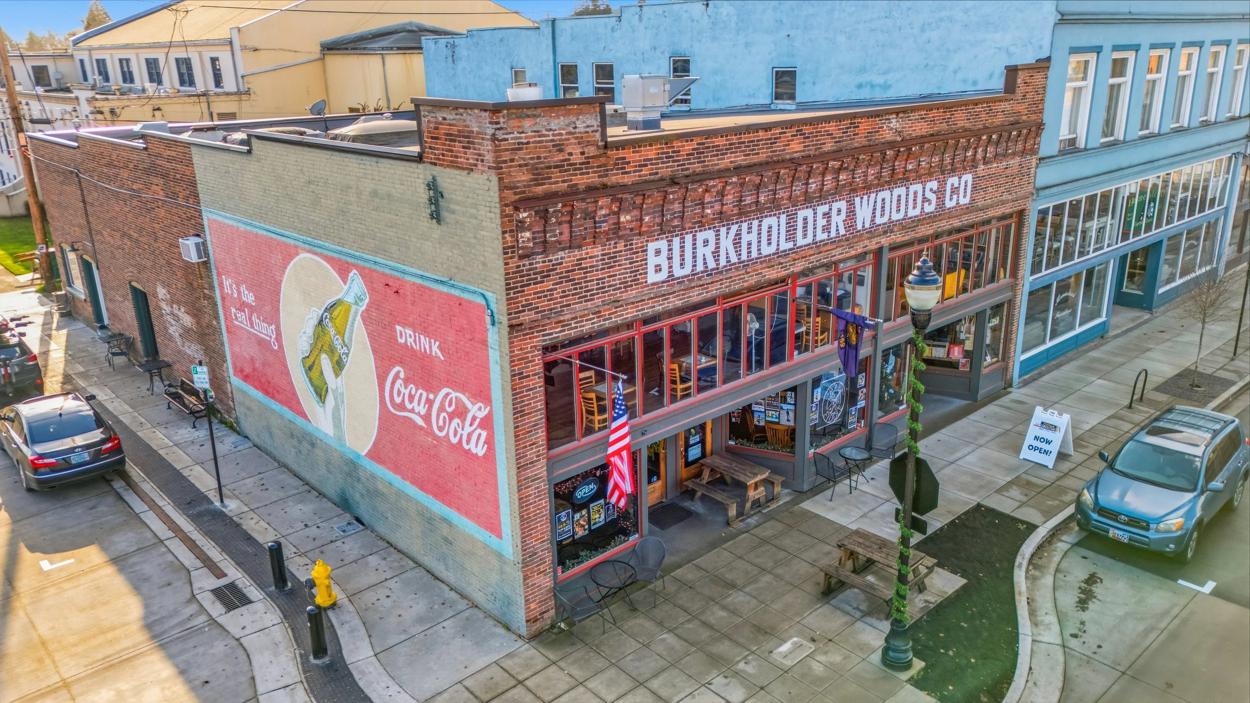 A brick building with a large painted Coca-Cola advertisement on the side wall, alongside a sign for Burkholderder Woods Co storefront with outdoor seating, American flags, and a sidewalk with a streetlamp and parked cars.