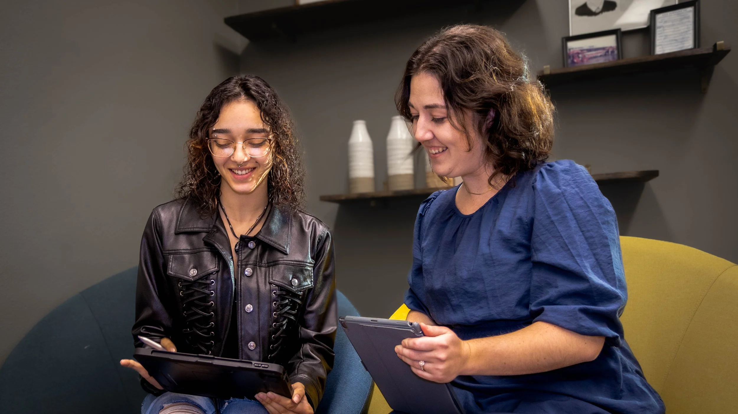 Two women looking at tablets, sitting on a yellow and blue sofa in a cozy indoor setting with framed pictures and decorative items on black shelves in the background.