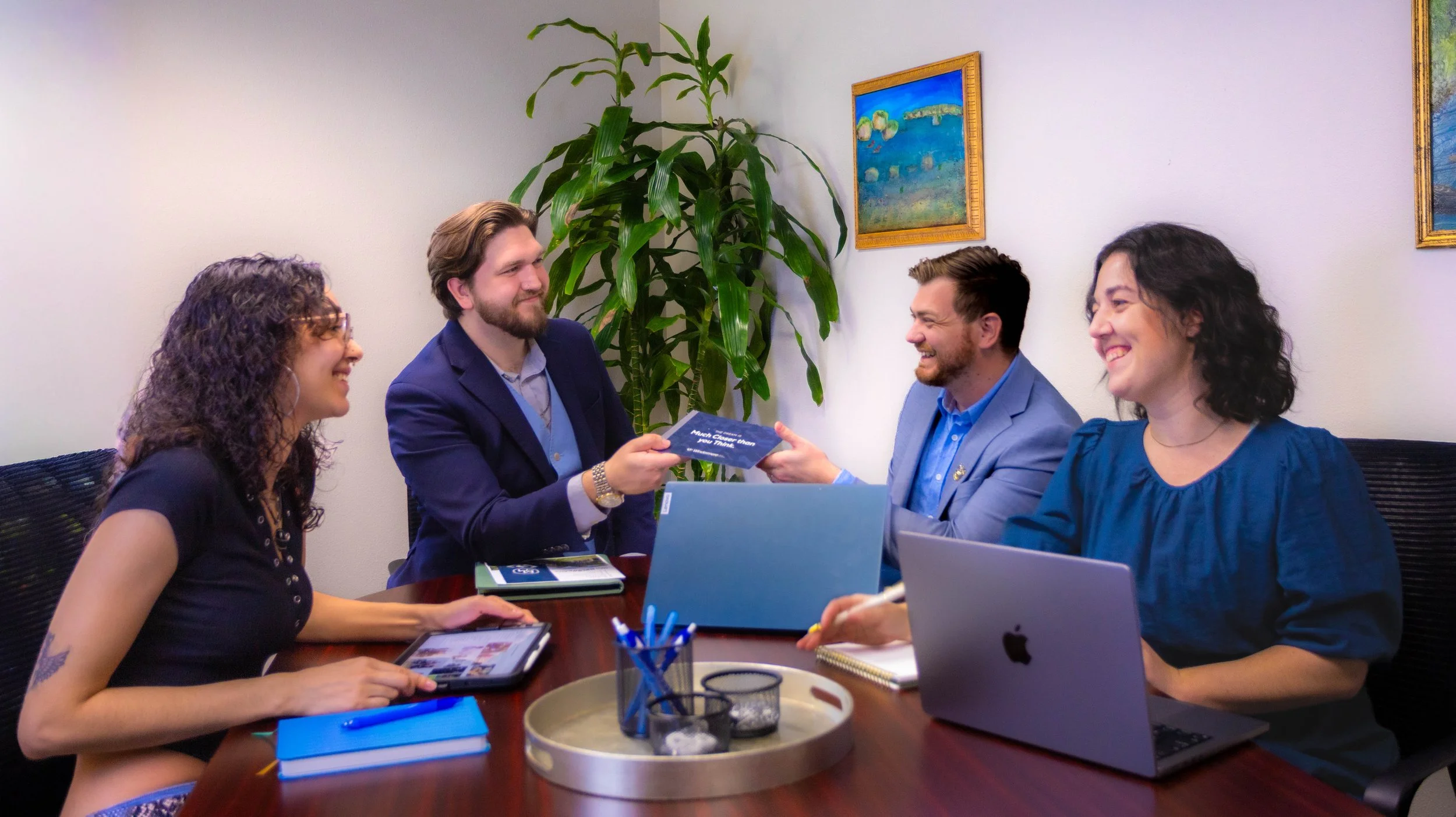 Four diverse colleagues sitting around a conference table, exchanging smiling and shaking hands, with laptops, notebooks, and pens in a modern office space