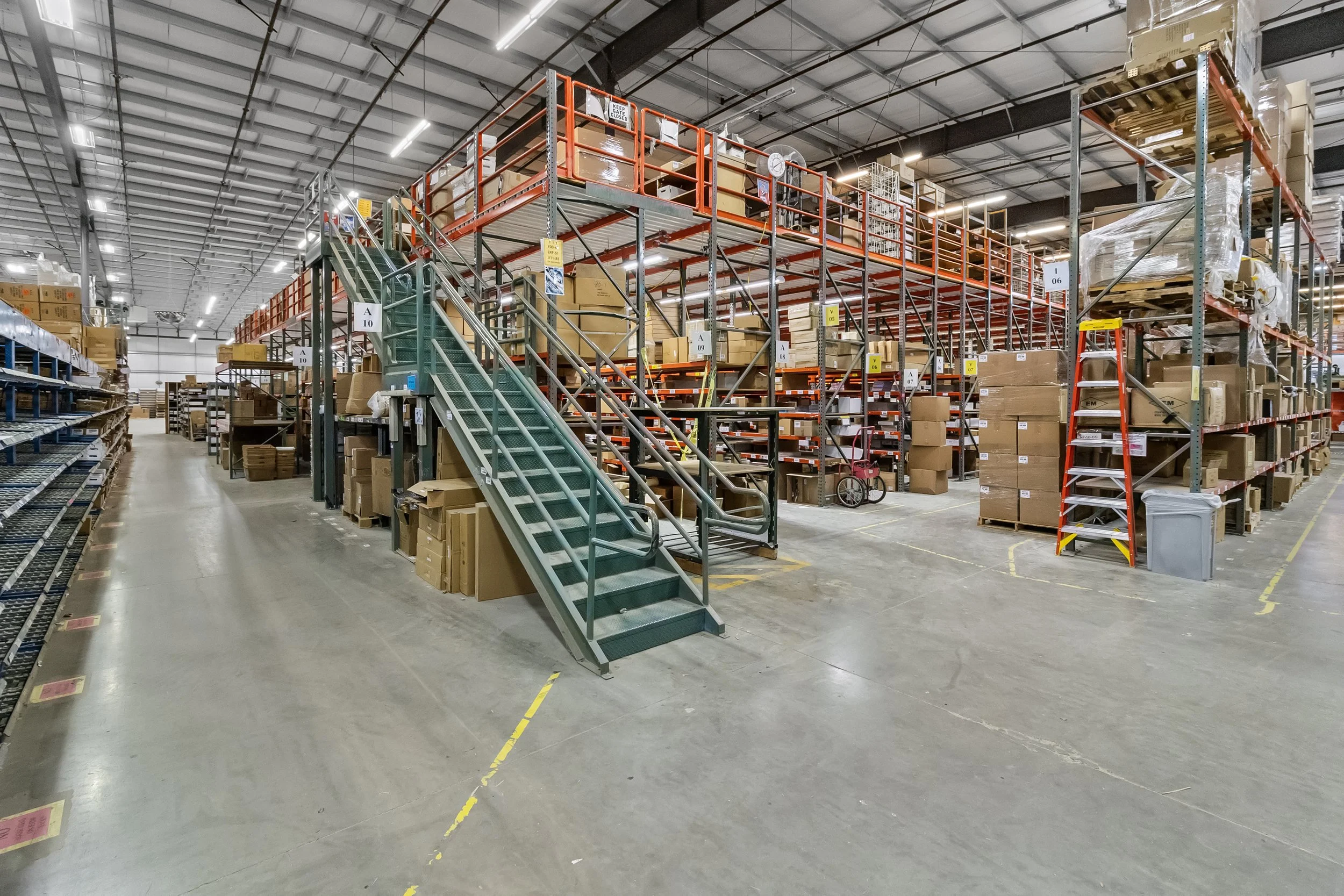 Inside a large warehouse or storage facility with metal shelving, cardboard boxes, and a staircase leading to an upper level of storage.
