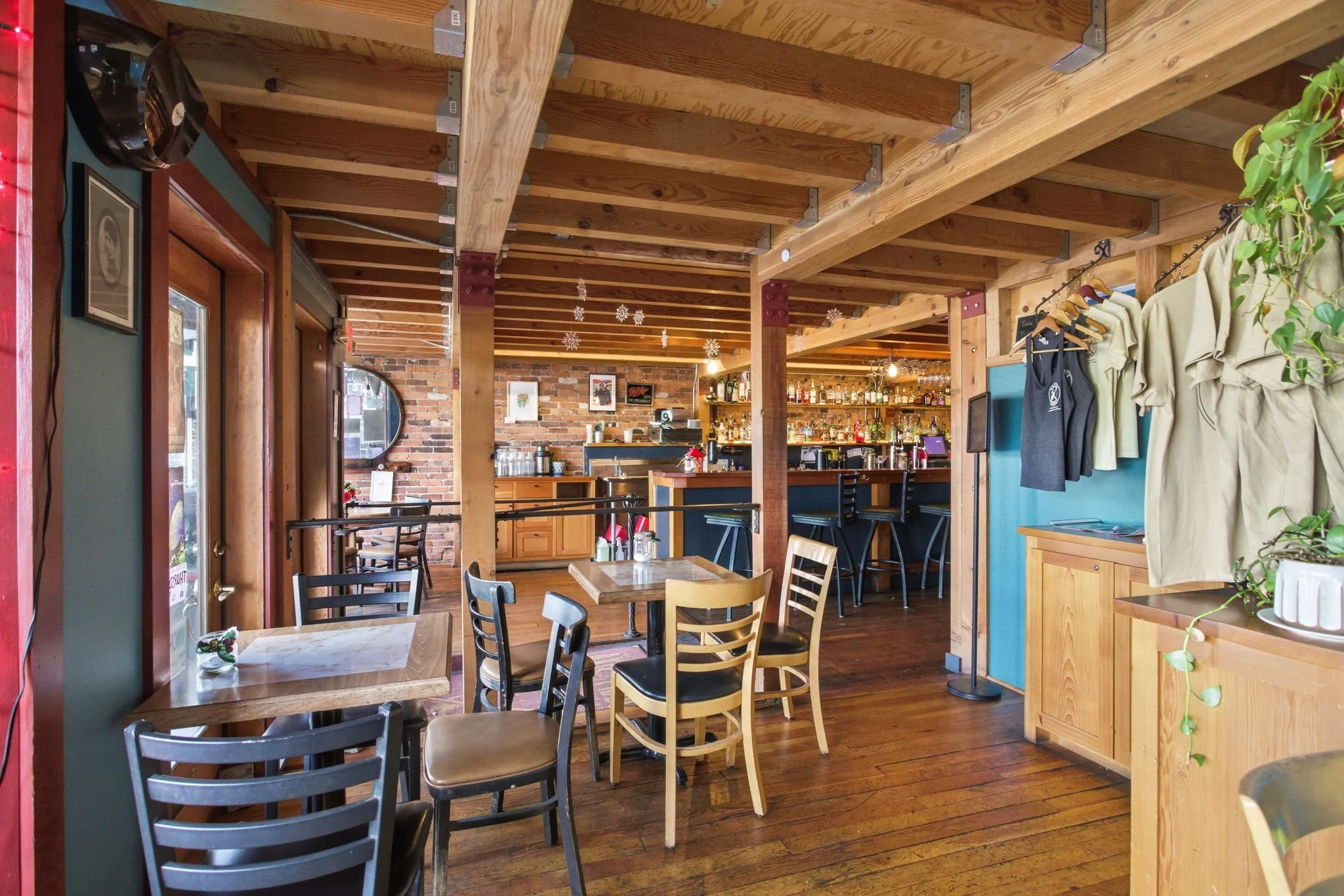 Interior of a cozy restaurant or café with wooden floors and walls, featuring tables and chairs, a bar with liquor bottles, and decorative snowflakes hanging from the ceiling.