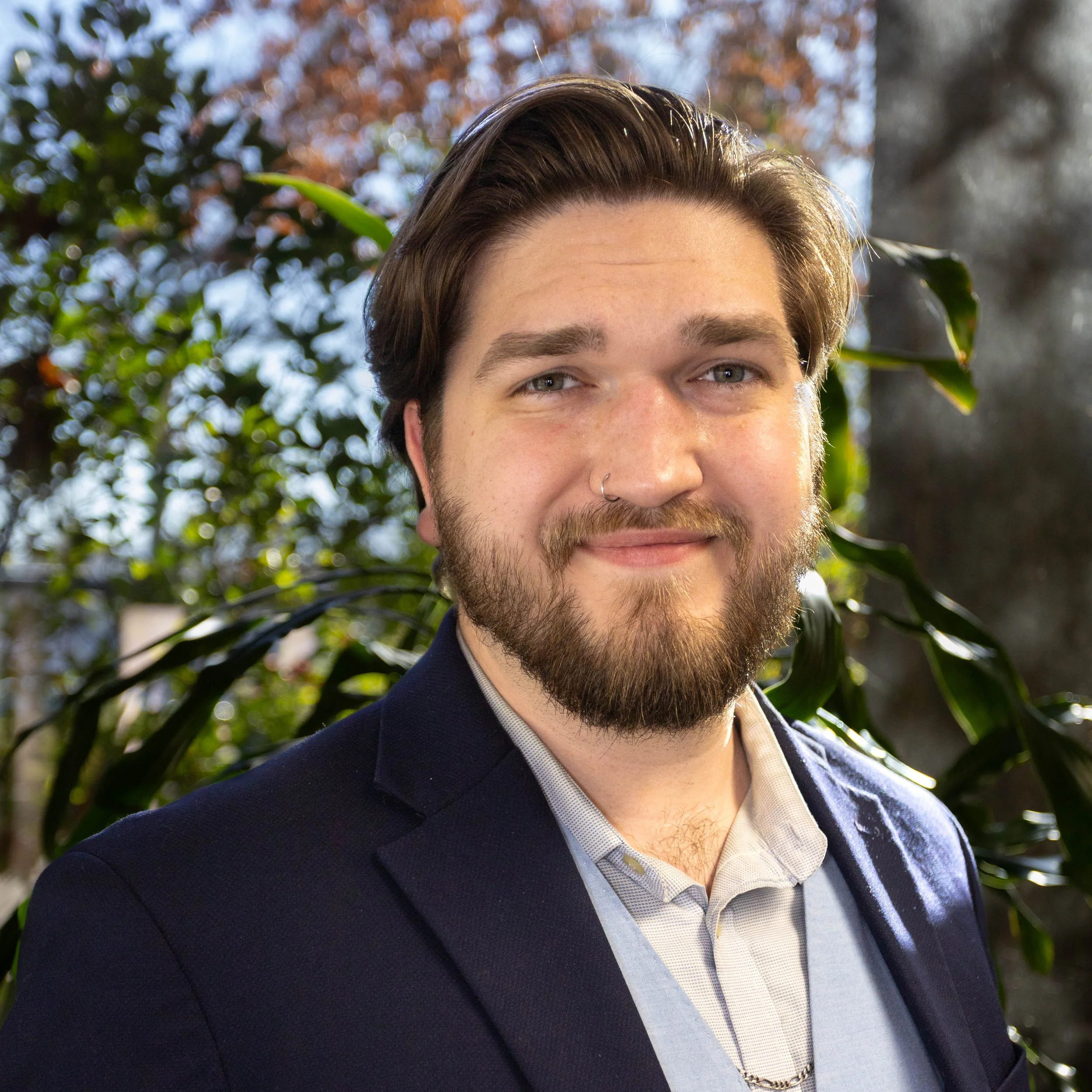 A confident young man with a beard, wearing a blue blazer and light-colored shirt, smiling in front of foliage with trees and leaves.