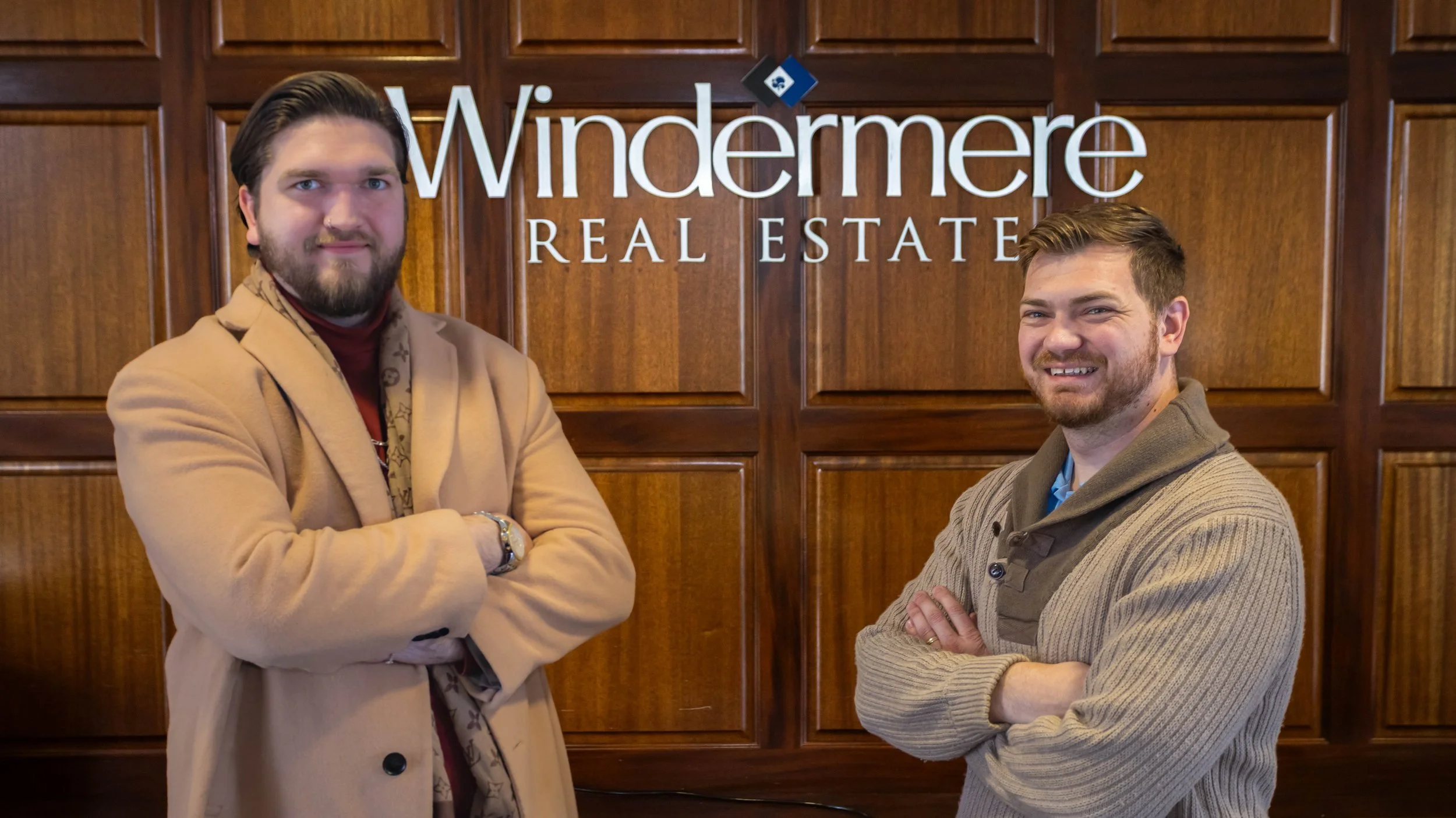 Two men standing in front of a wooden wall with a sign that reads 'Windermere Real Estate'. Both men are smiling and have their arms crossed.