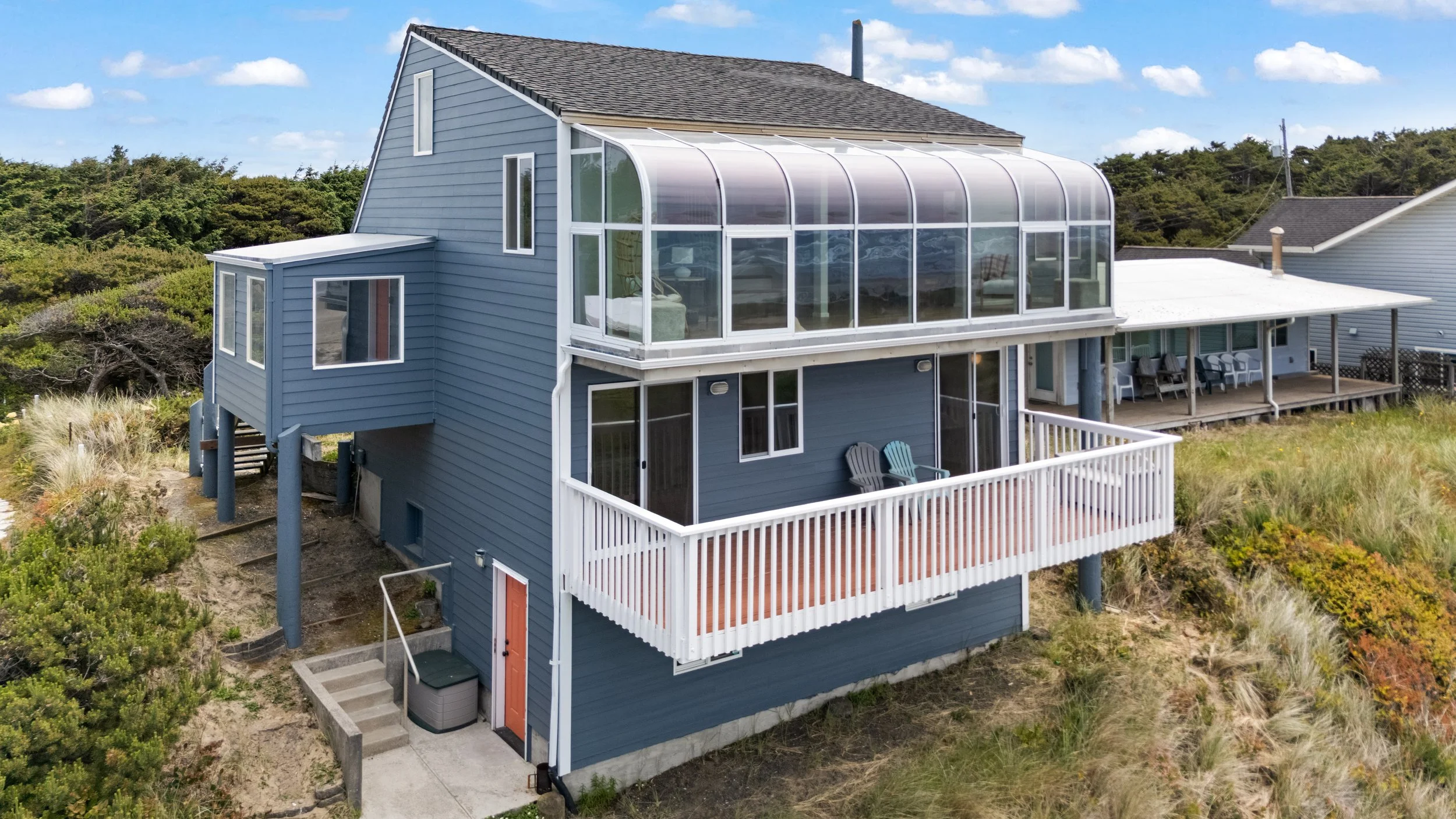A blue multi-story house with a large enclosed balcony or sunroom on the upper level, outdoor balcony on the lower level with chairs, in a coastal or beachside environment with grassy dunes and vegetation around.