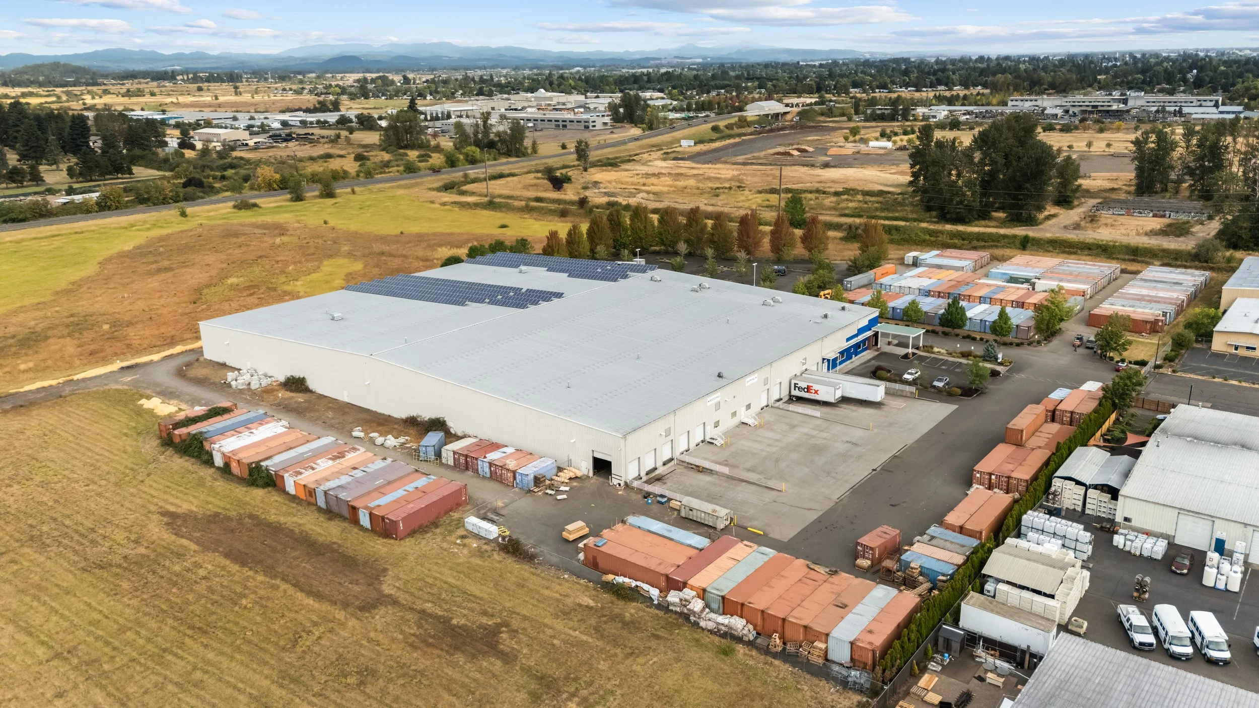 Aerial view of a large industrial warehouse with a FedEx truck parked outside, surrounded by shipping containers and parked cars, with fields and other buildings in the background.